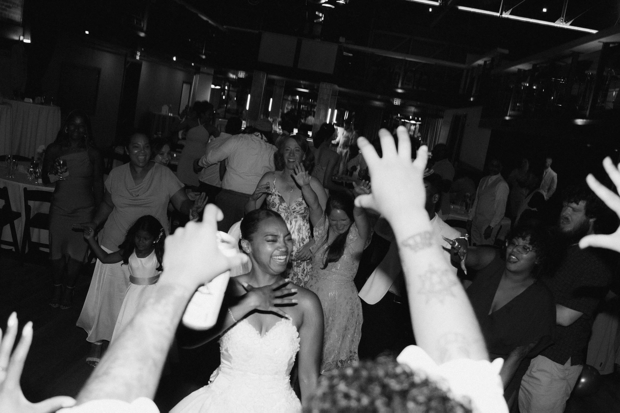 Group of people dancing and celebrating at a wedding reception with a bride in a white dress smiling in the center.