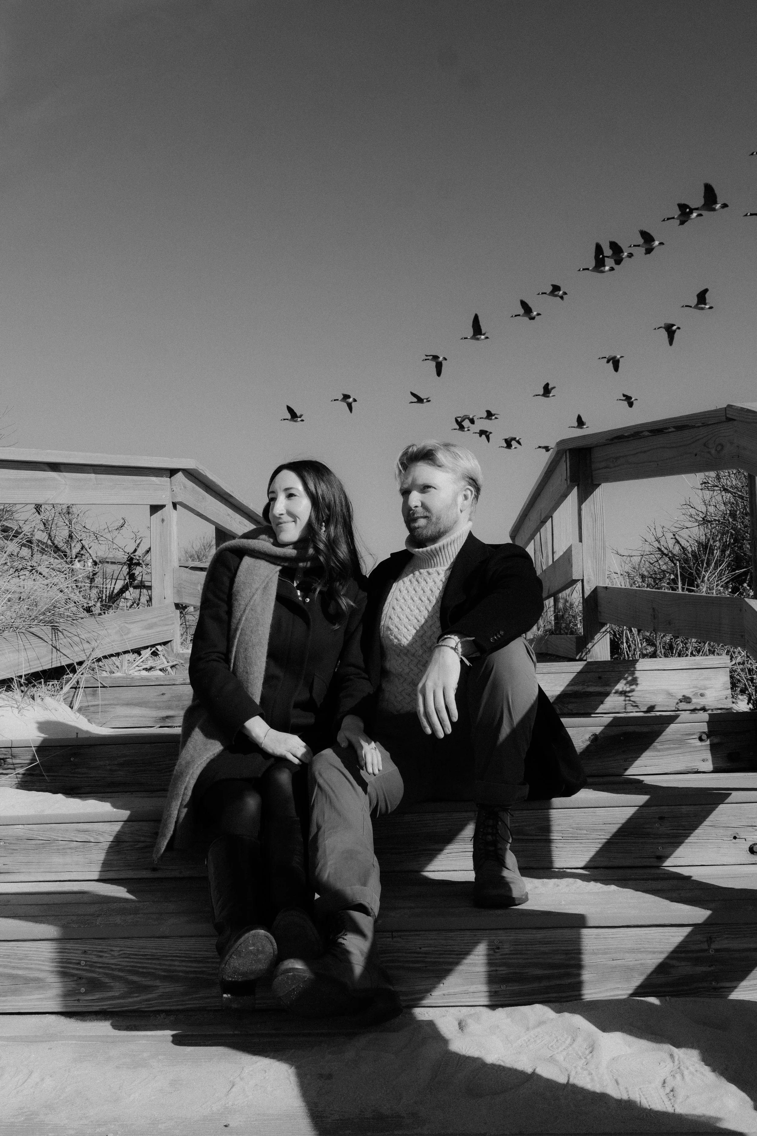 Black and white photo of a man and woman sitting on wooden steps outdoors, with a flock of birds flying in the sky above them.