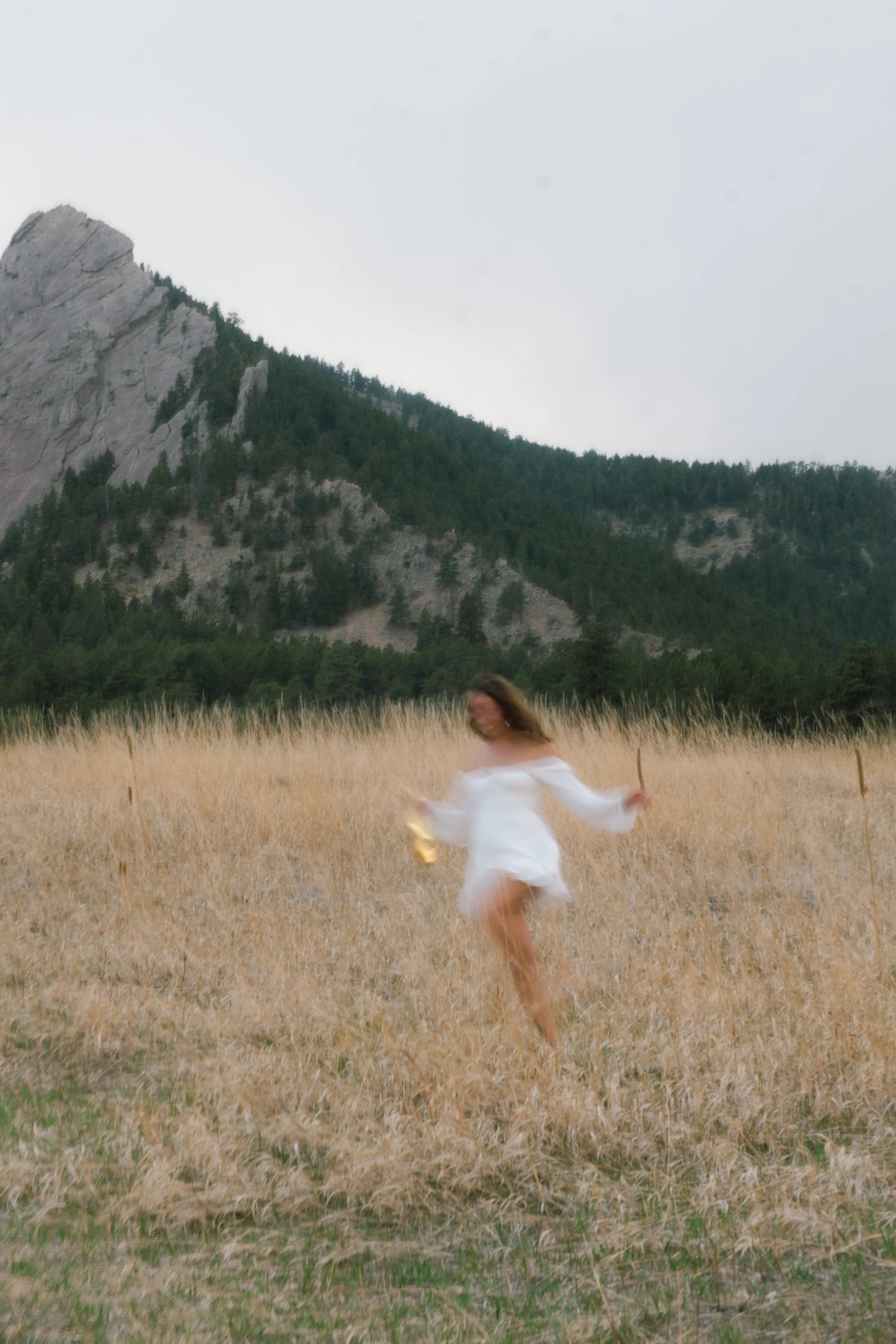 A woman in a white dress is dancing in a field of tall, golden grass, with mountains and trees in the background on a cloudy day.
