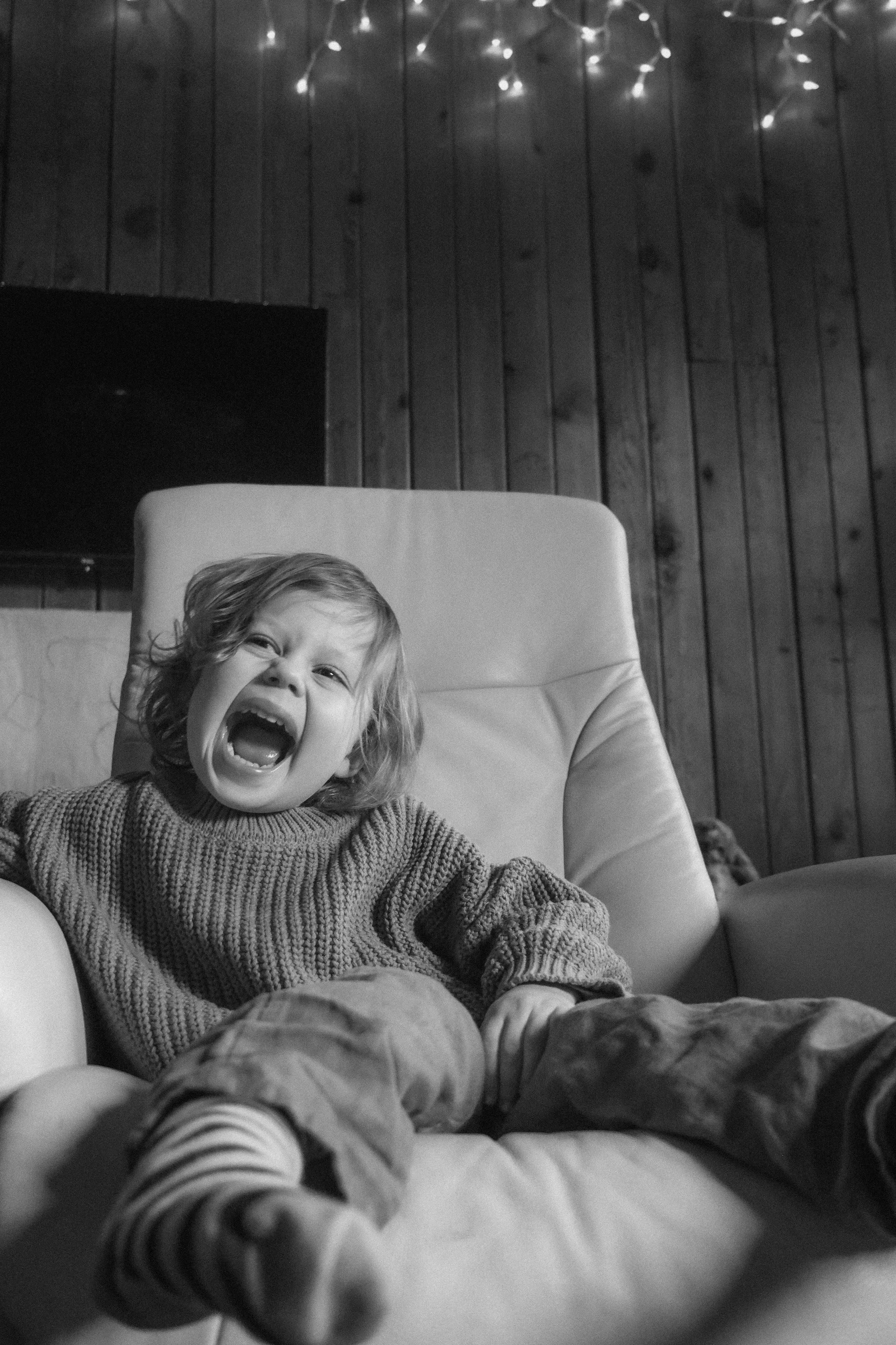 A young child with curly hair sitting on a large armchair, laughing or shouting with an open mouth, in a room with wooden paneling and string lights hanging from the ceiling.