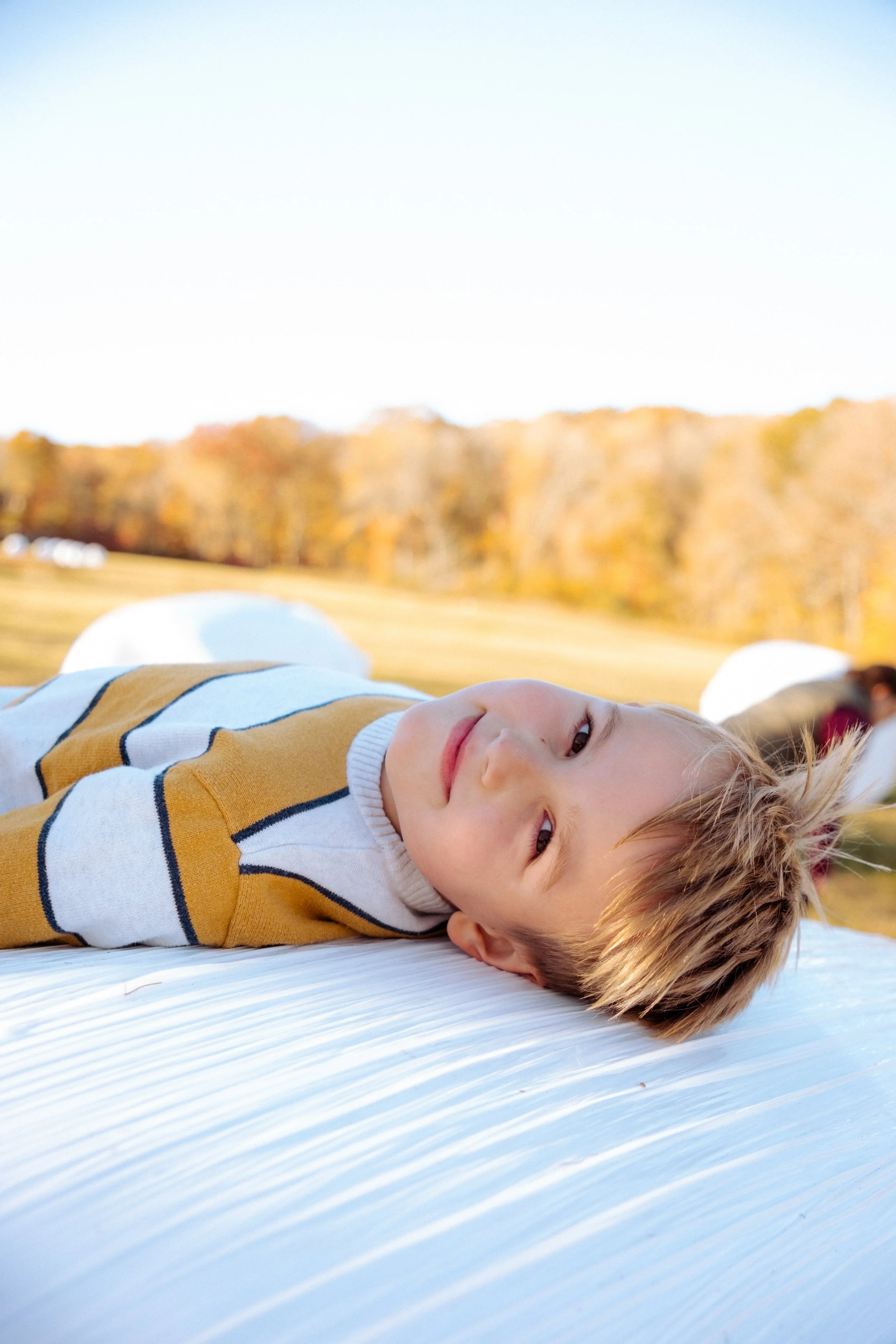 A young boy with blond hair smiling, lying down on a white surface outdoors during fall, with trees in the background.