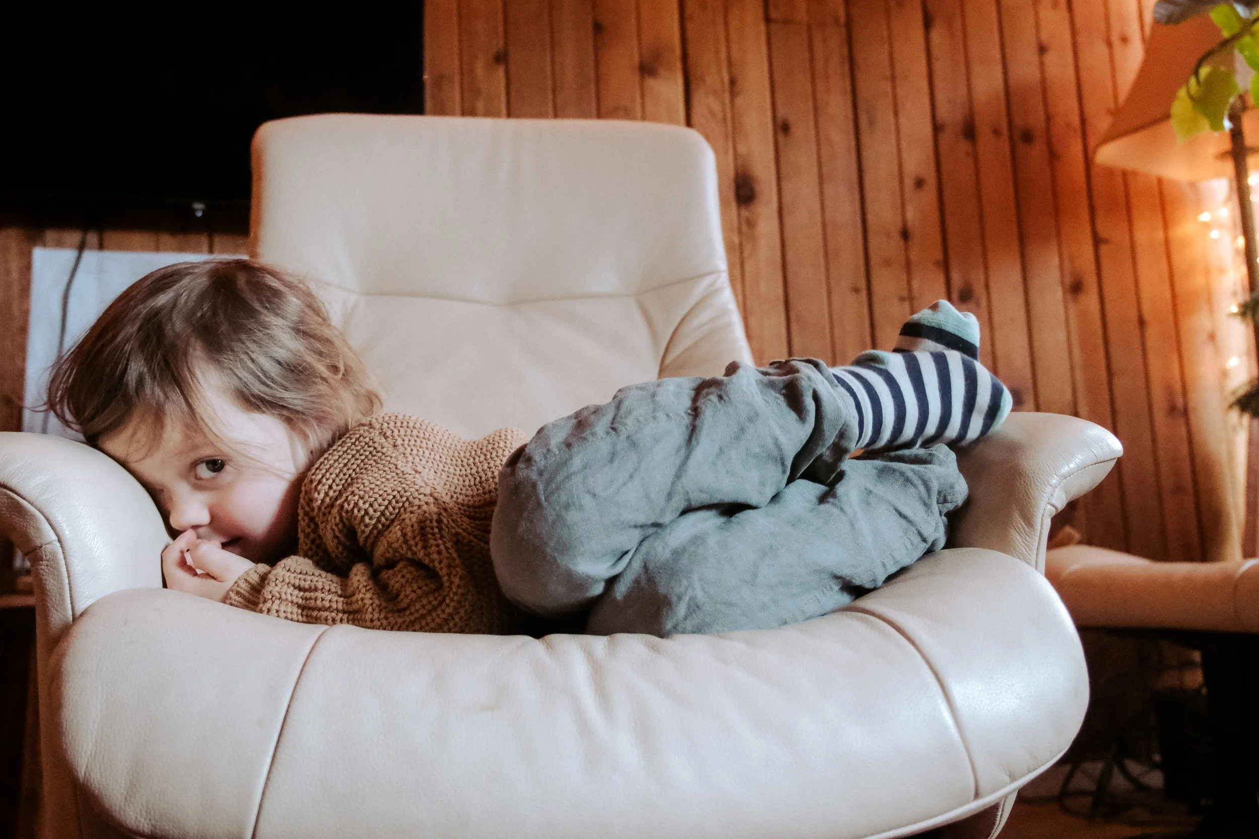 A young child with brown hair and a brown sweater lying on a cream-colored armchair, curled up and smiling at the camera with a cozy wooden wall and a partially visible chair with a yellow cushion in the background.