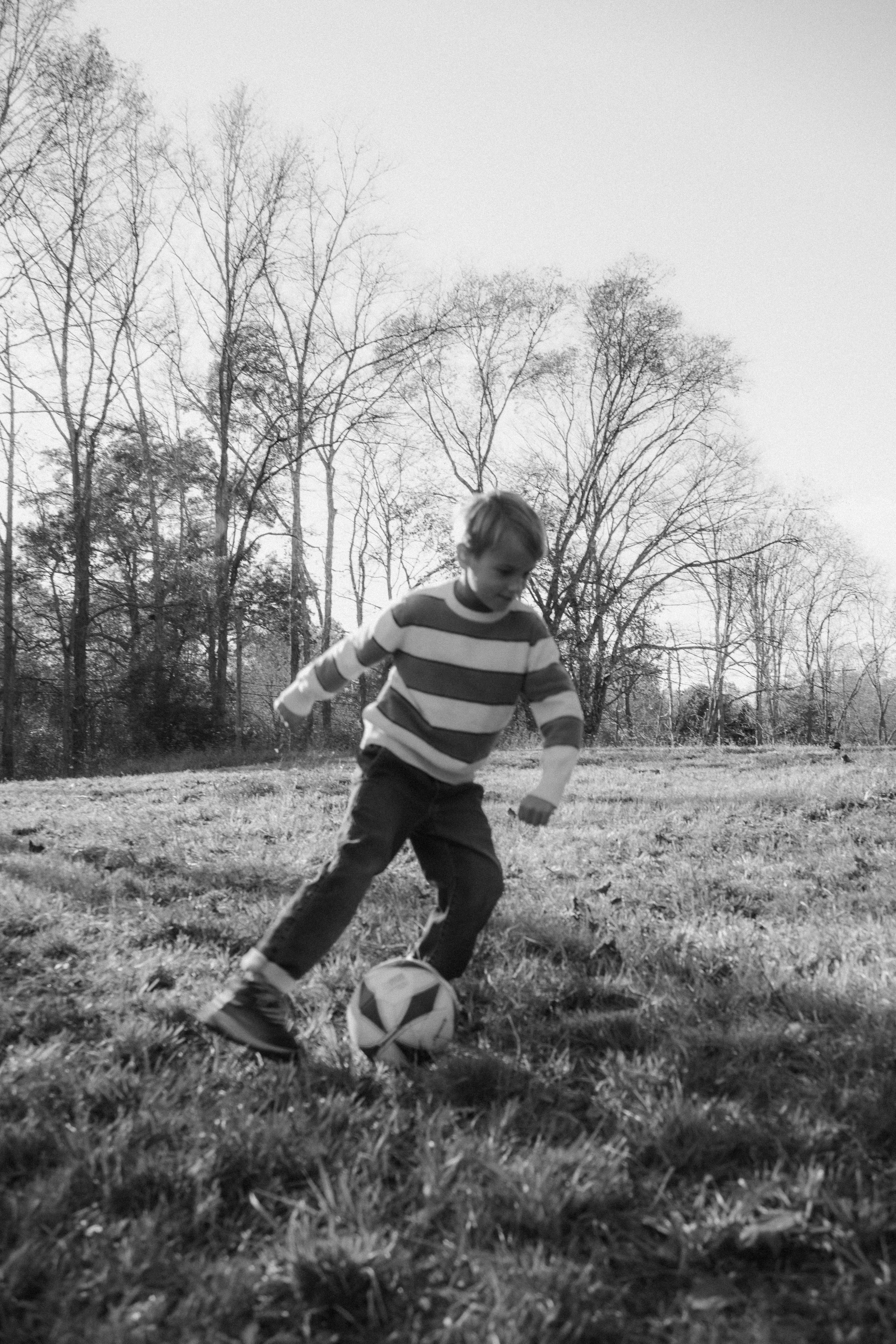 A young boy wearing a striped long-sleeve shirt and dark pants playing soccer outdoors on a grassy field surrounded by leafless trees, captured in black and white.