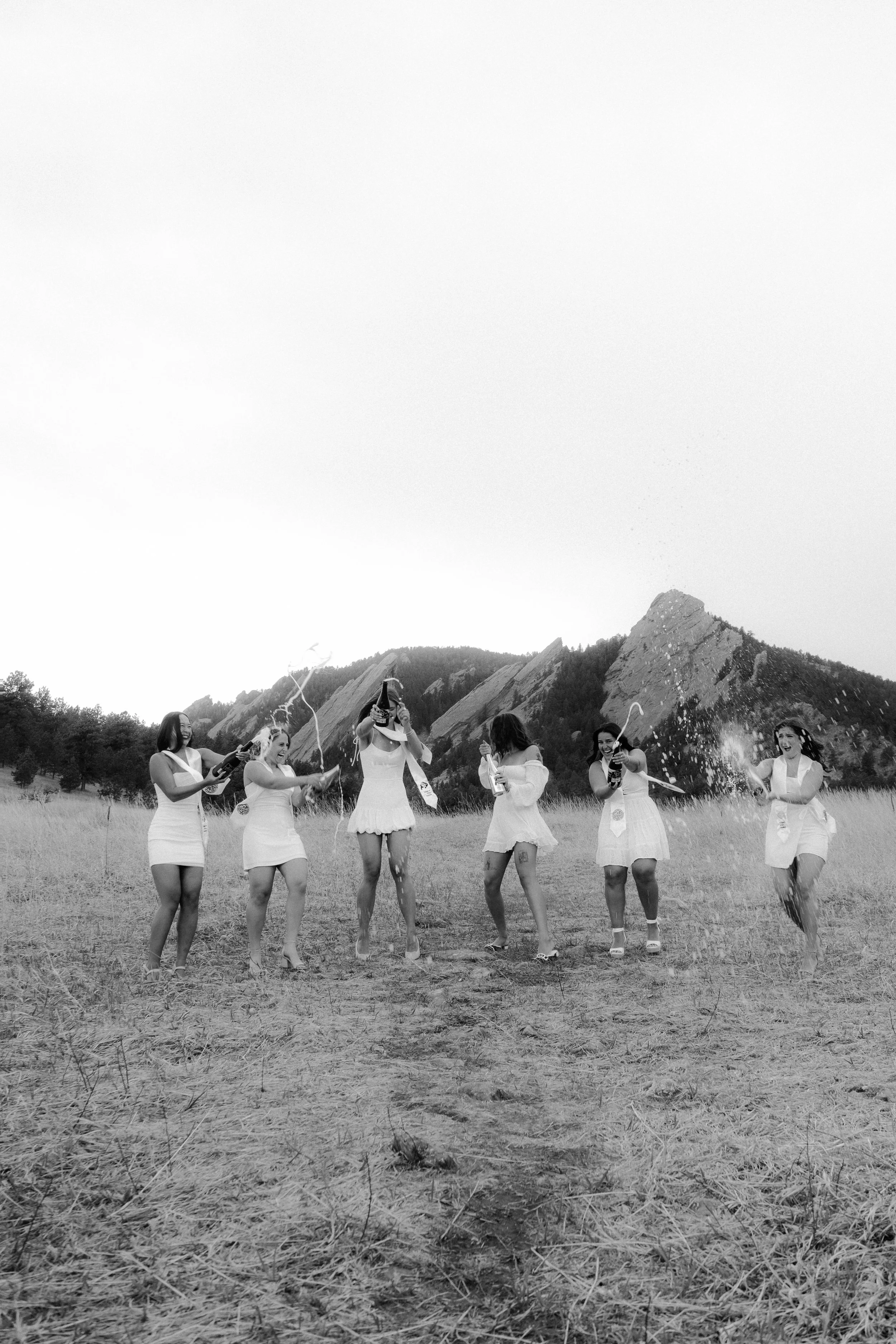 Seven women in white dresses celebrating with party poppers in an open field, mountains in the background, black and white photo.