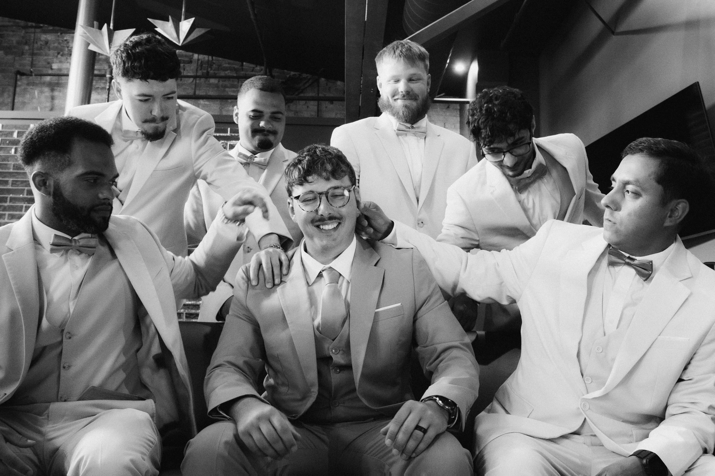 Group of men dressed in formal suits and bow ties, with some smiling and touching each other's shoulders, indoors with brick wall and modern decor.