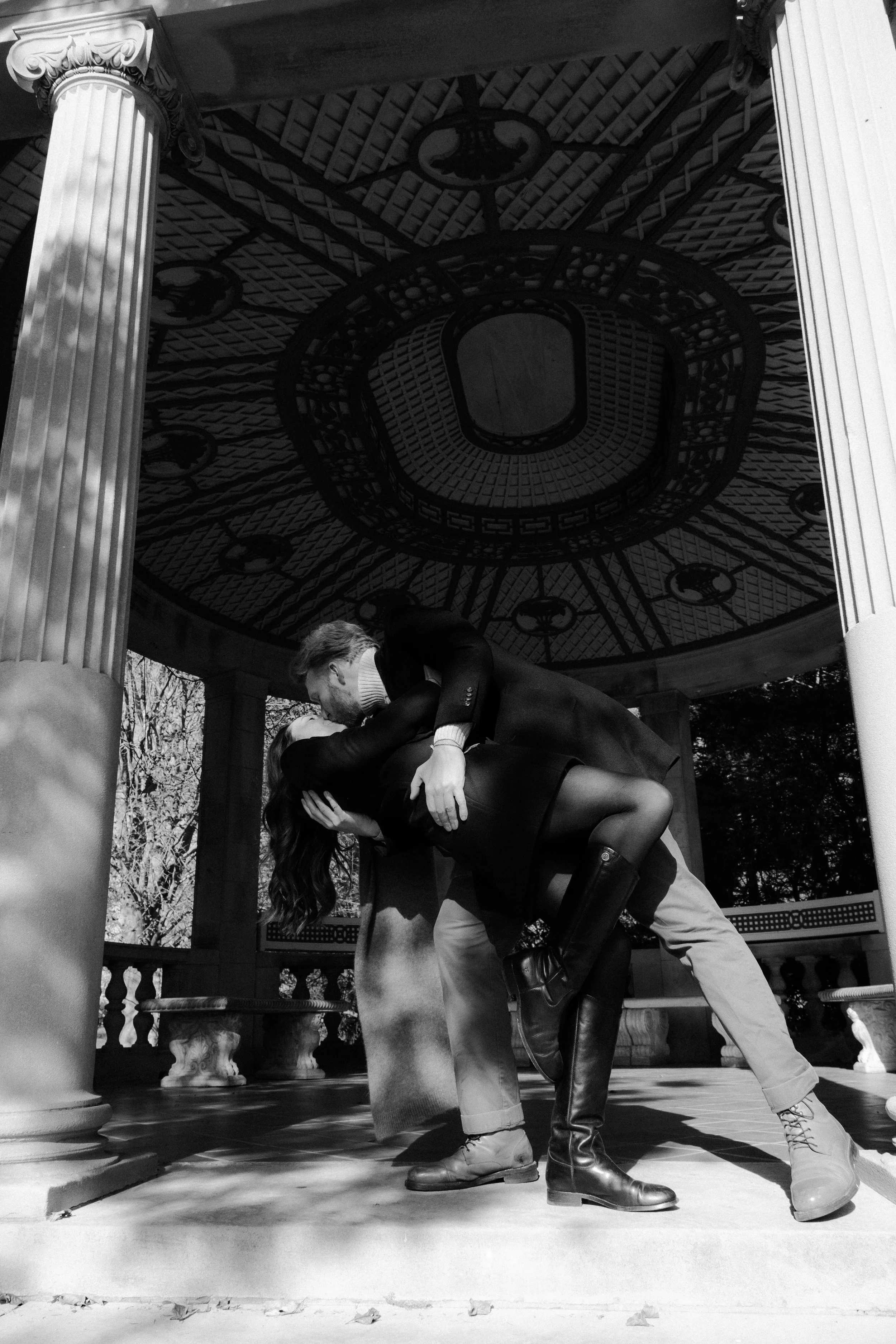 A black and white photo of a couple dancing closely under a gazebo with ornate columns, with one person dipping the other. The gazebo has a decorative circular ceiling with Roman numerals, and sunlight creates shadows on the floor.