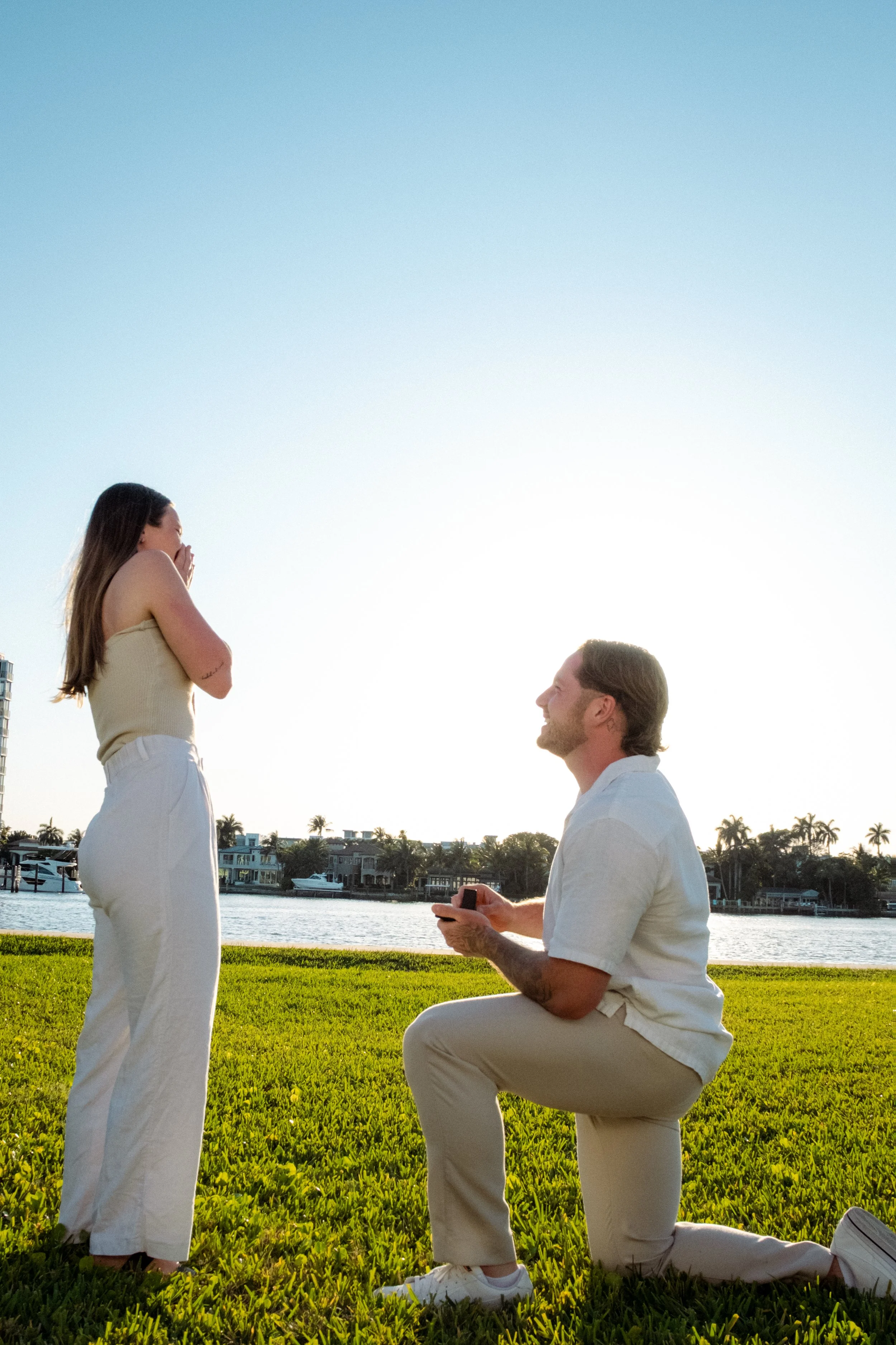 A man proposing marriage to a woman outdoors by a waterway during sunset, with the man on one knee holding a ring box and the woman covering her mouth in surprise.