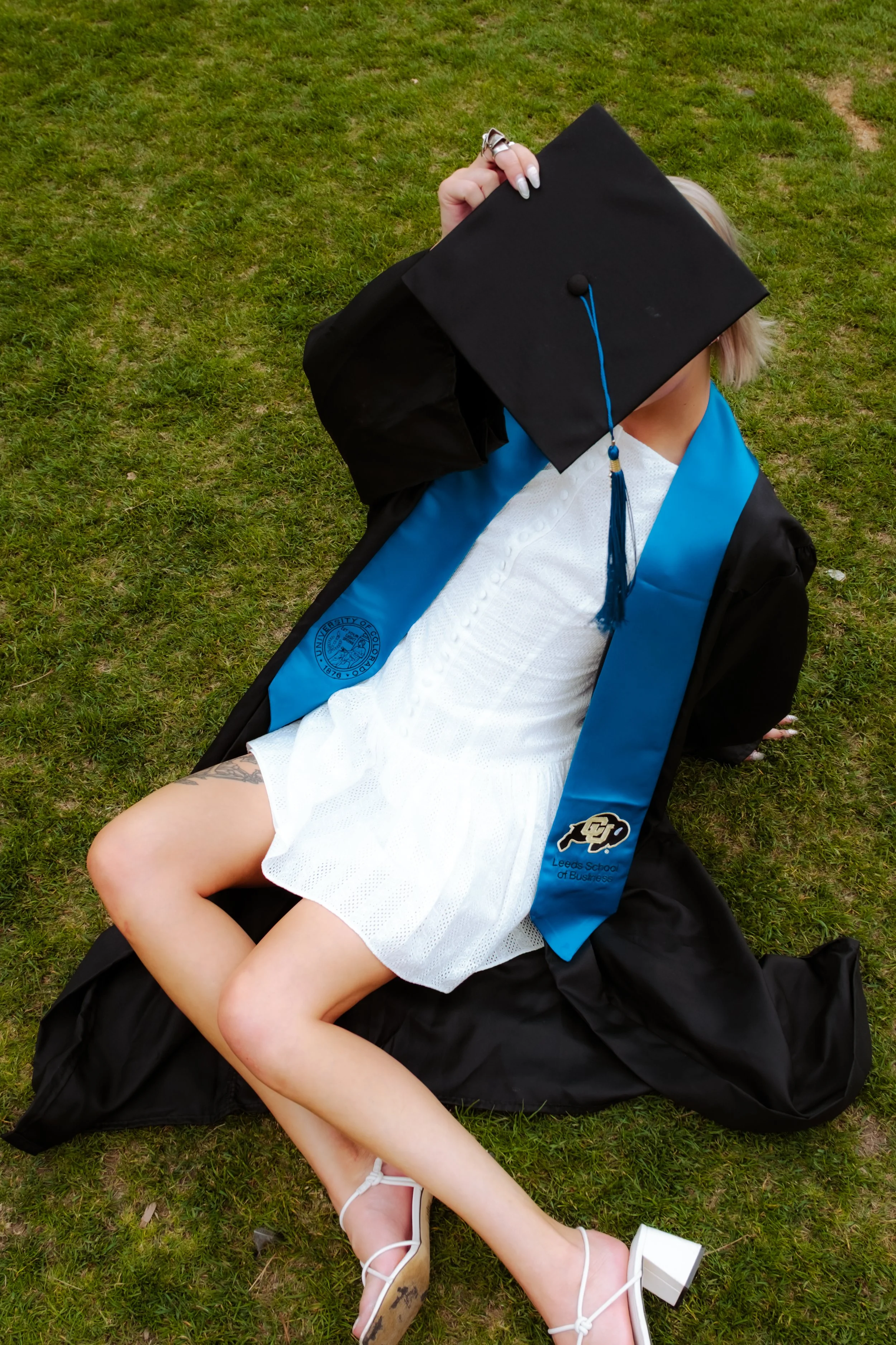 A person in a white dress and white heels sitting on grass with a graduation cap and gown, holding their face with the cap.