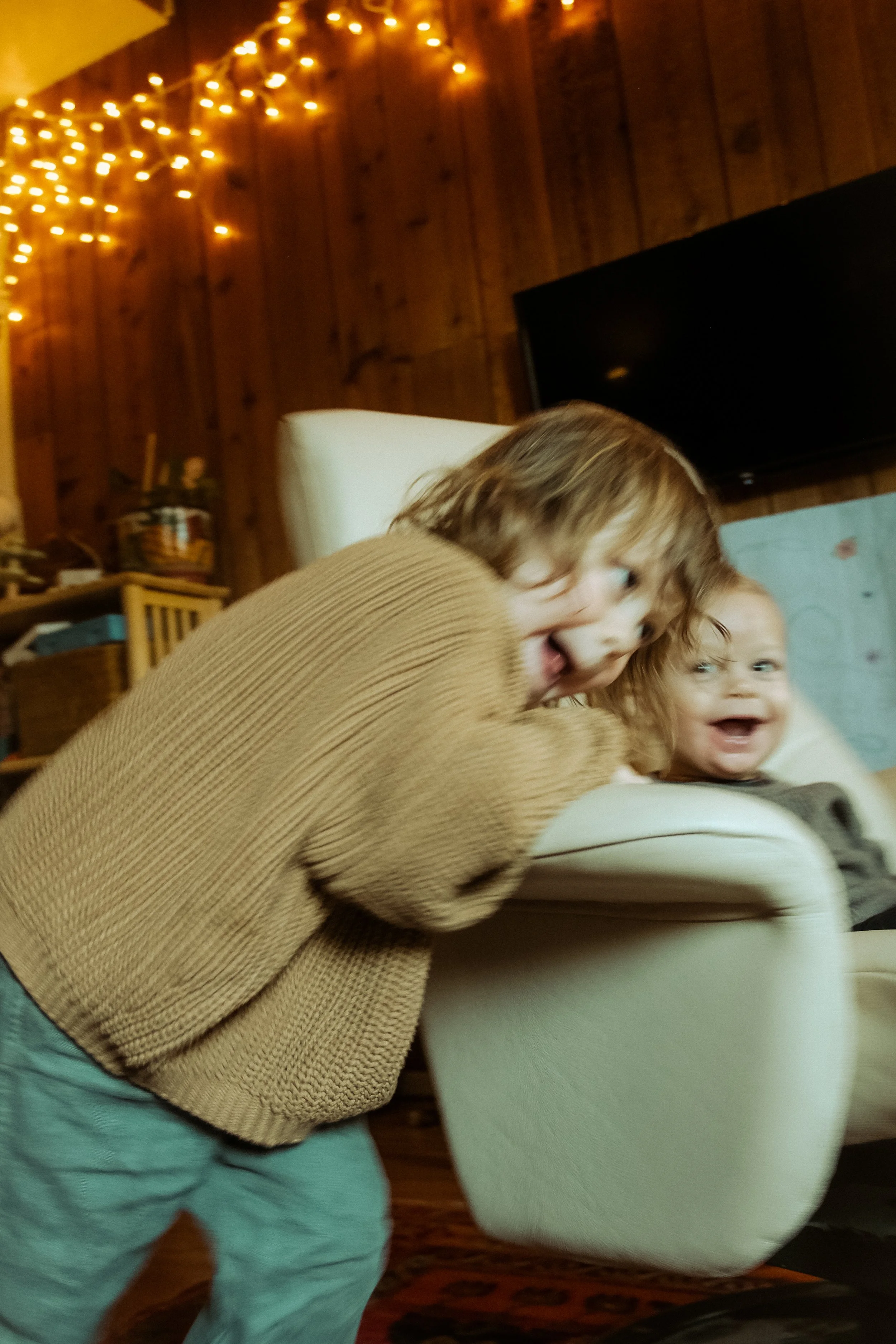 Two young children playing and laughing indoors, one leaning on a cream-colored armchair in a cozy room with string lights and wooden walls.