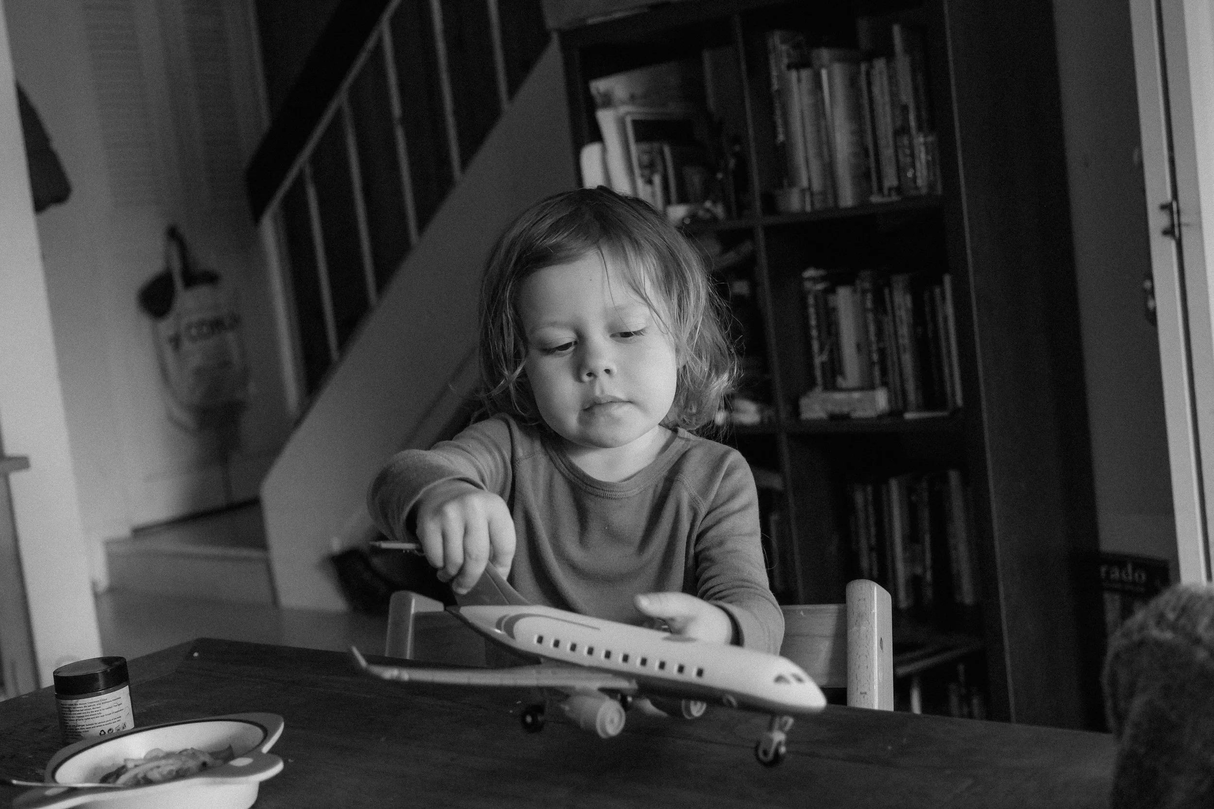 A young child playing with a toy airplane at a table in a living room, with bookshelves and a staircase in the background.