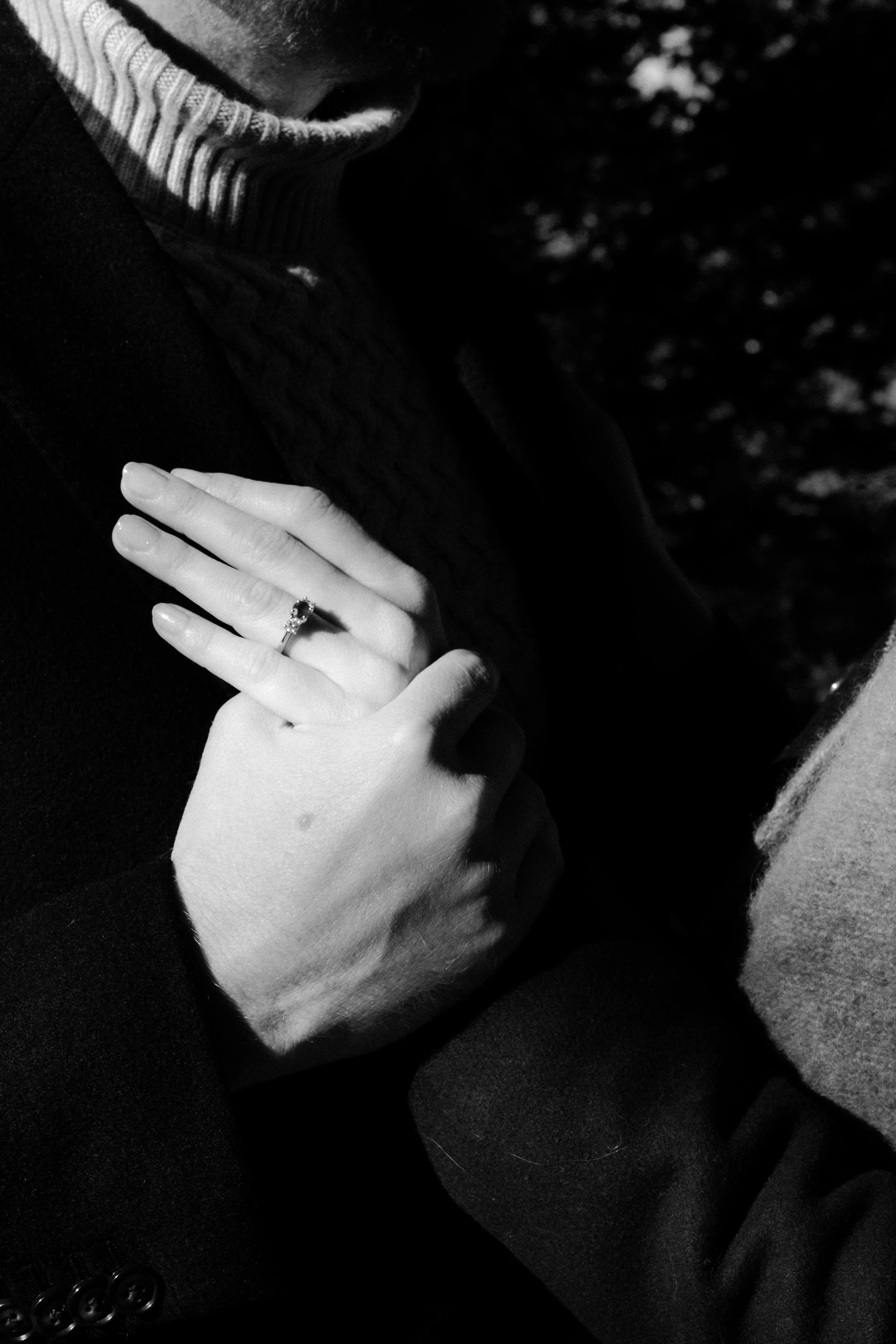 Close-up of a woman's hand with a ring, resting on a man's shoulder. Both are wearing dark clothing, with the woman's hand prominently displayed.