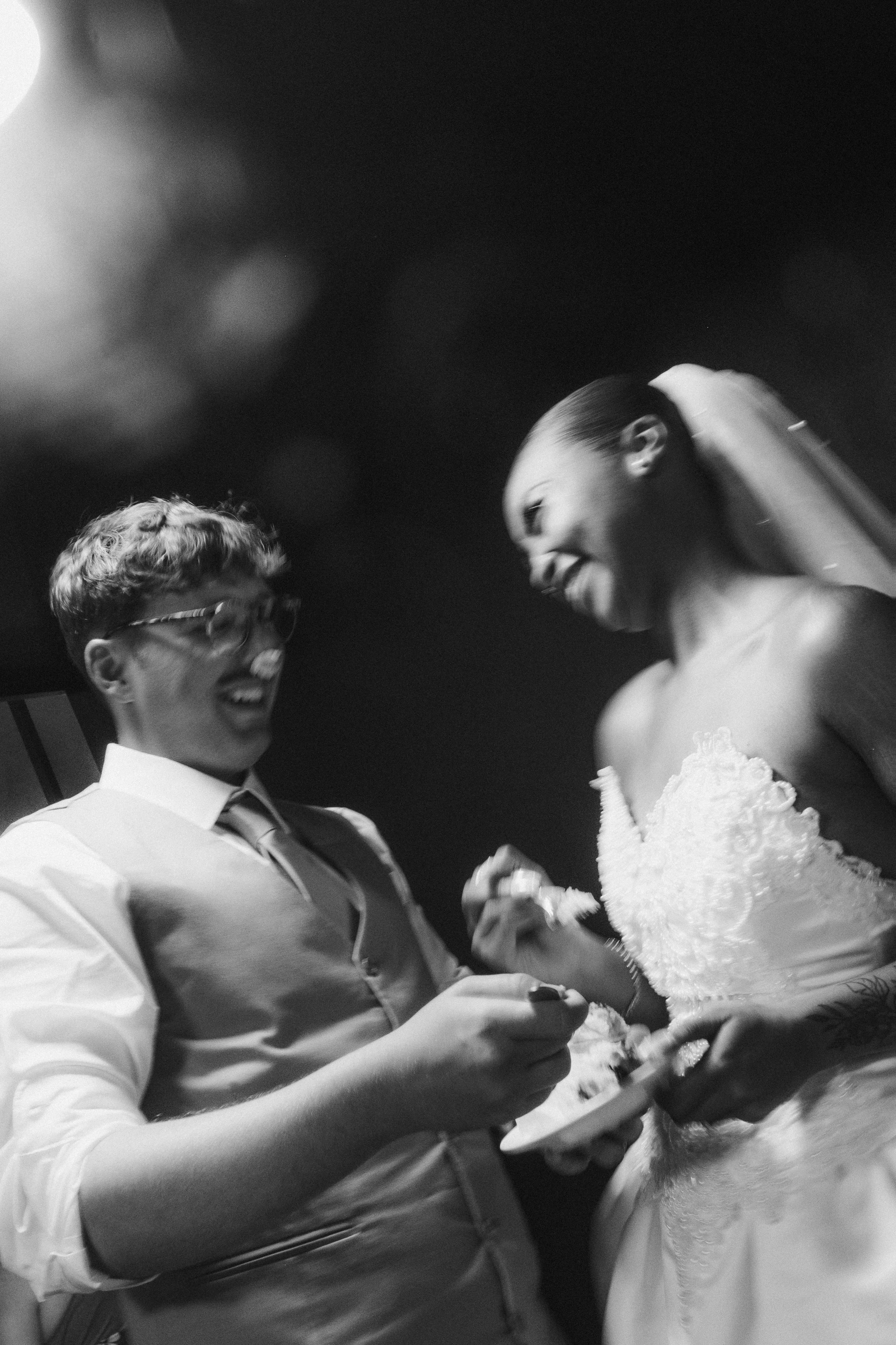 A black-and-white photo of a couple laughing and sharing a moment at their wedding. The man is wearing glasses and a vest, holding a piece of wedding cake, while the woman is in a wedding dress with a veil, holding a plate and a fork.