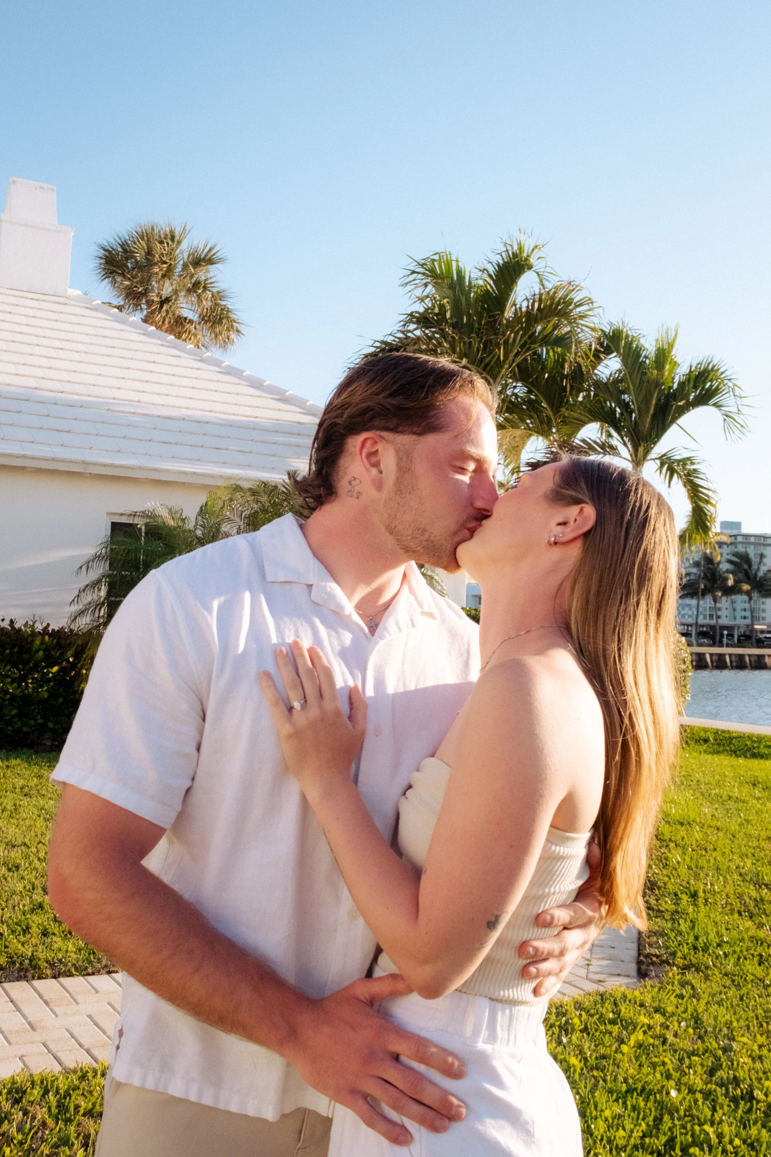 A couple sharing a kiss outdoors, with palm trees and a building in the background, during daytime.