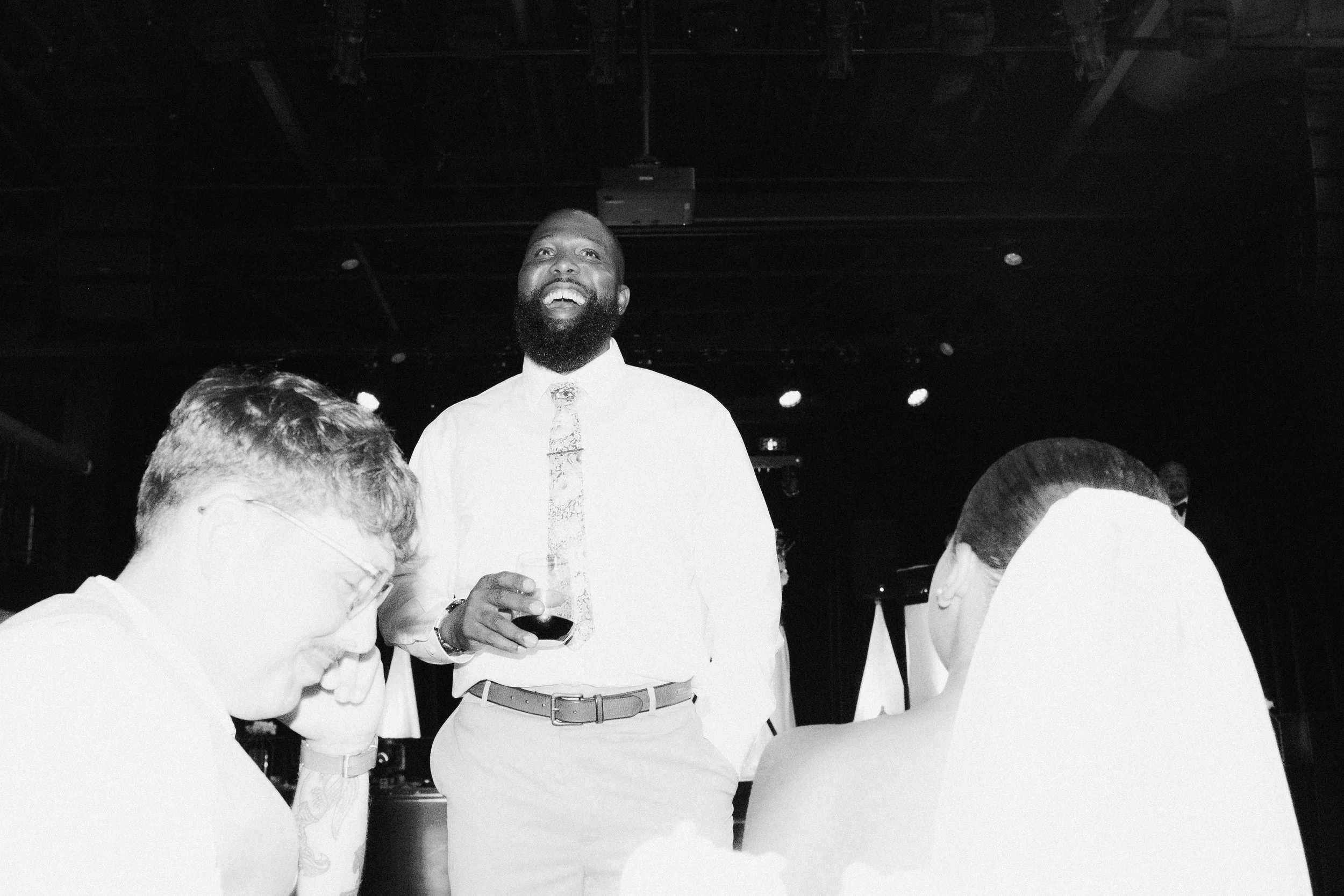 A man with a beard in a dress shirt and tie laughs while standing at a social gathering, with two other people with glasses and ponytail sitting in the foreground.