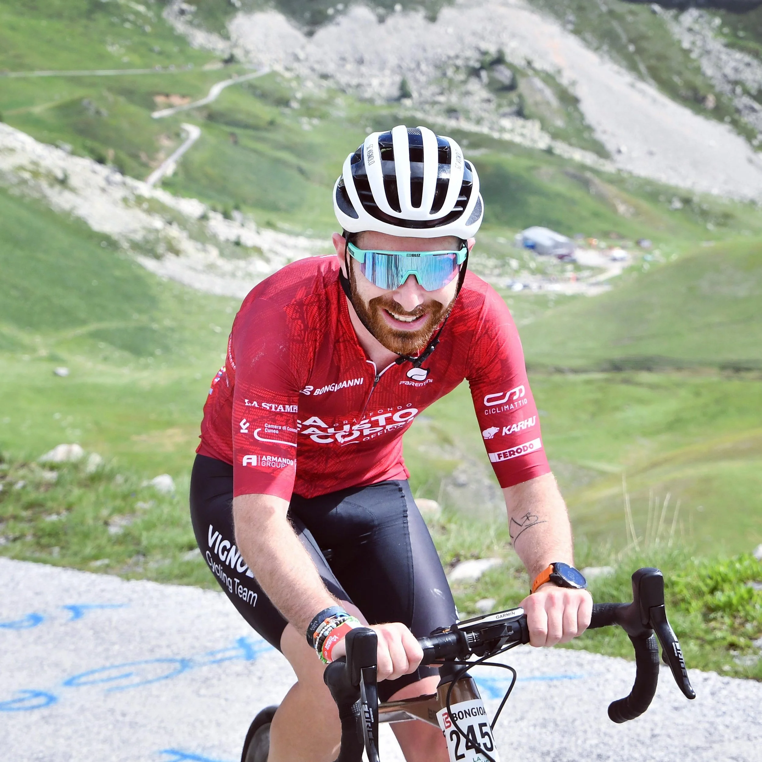 Un uomo sorridente seduto su una roccia sulla spiaggia, indossa una maglietta grigia, pantaloncini neri, orologio e ha uno zaino sulla schiena, con il sole e il cielo azzurro sullo sfondo.