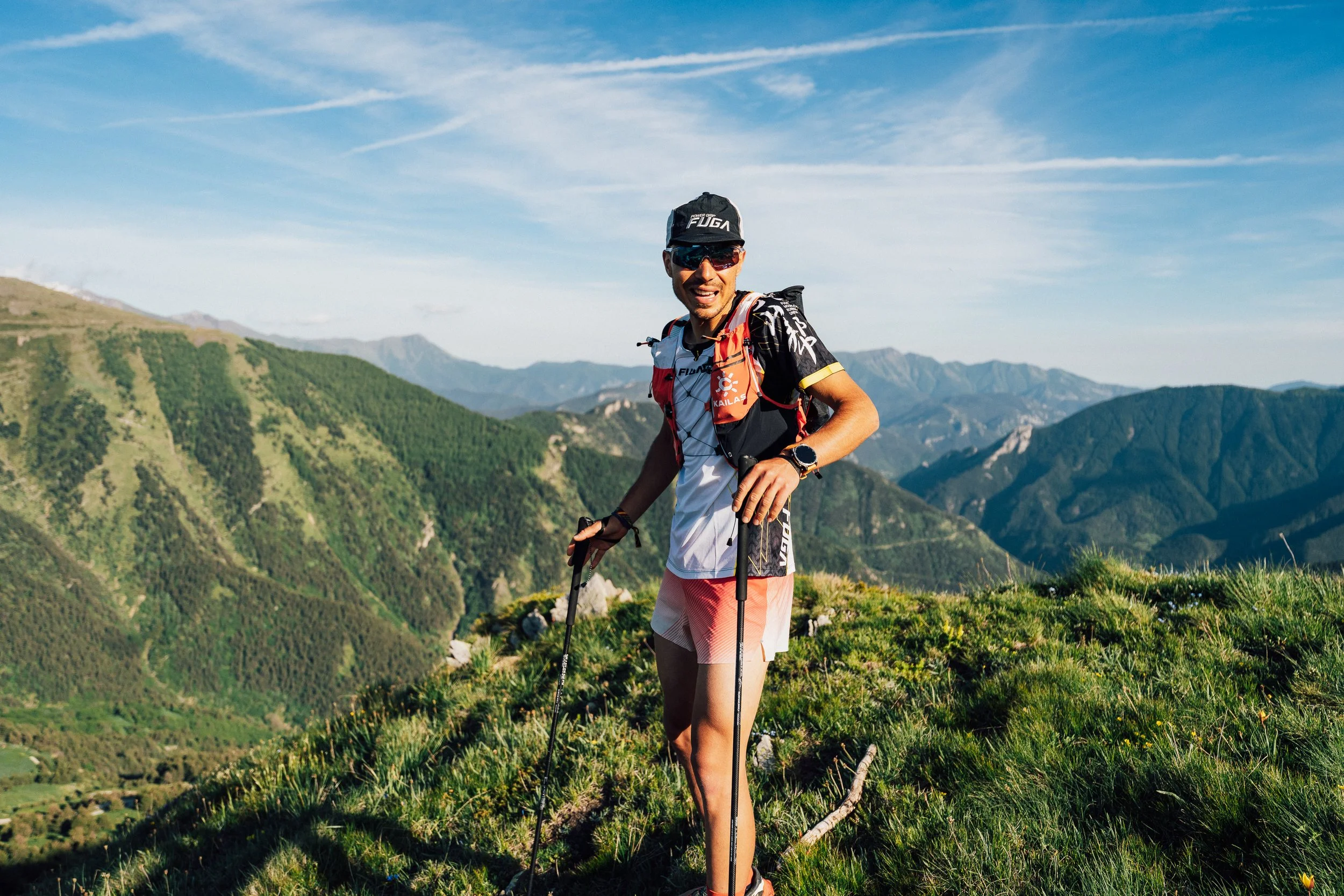 Ragazzo sorridente con occhiali, in maglietta blu, in un ambiente con molte piante verdi e una struttura in legno sullo sfondo.