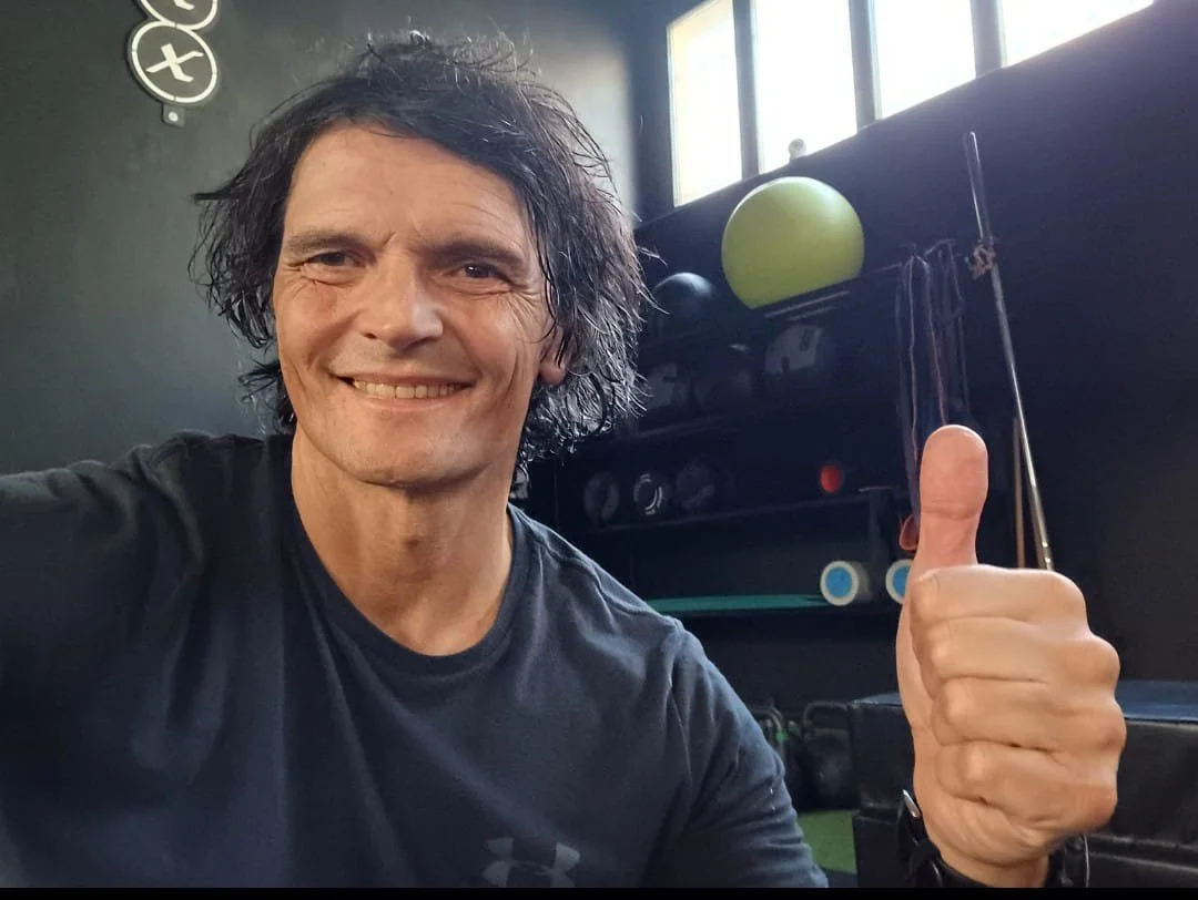 Ritratto di un uomo con capelli ricci, barba e occhiali tondi, che sorride, indossa una camicia con un motivo floreale e uno sfondo grigio chiaro.