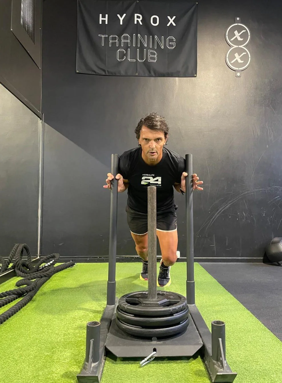 Ritratto di un uomo con capelli ricci, barba e occhiali tondi, che sorride, indossa una camicia con un motivo floreale e uno sfondo grigio chiaro.