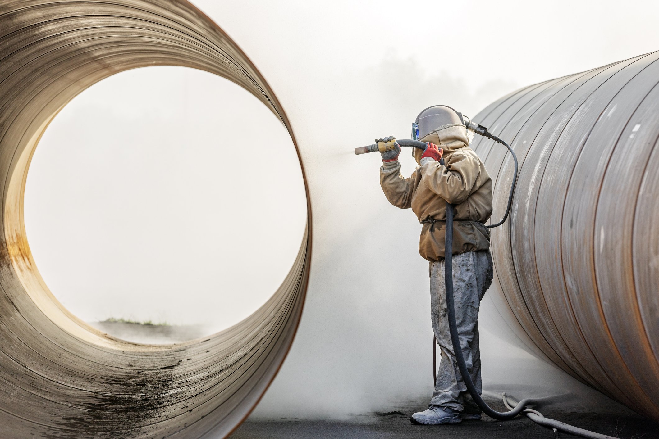 Worker wearing protective gear uses a welding tool on large metallic pipes in an industrial setting.