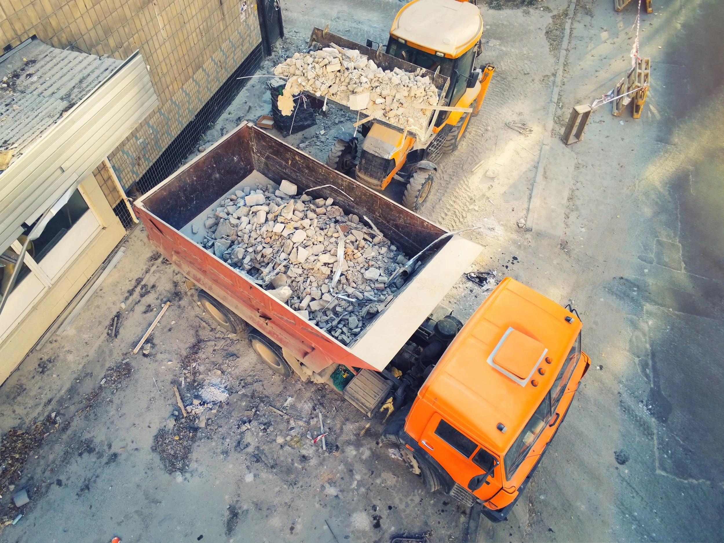 High-angle view of a construction site showing an orange dump truck filled with rocks and debris, a yellow loader dumping rocks into the truck, and scattered construction materials and dirt on the ground.