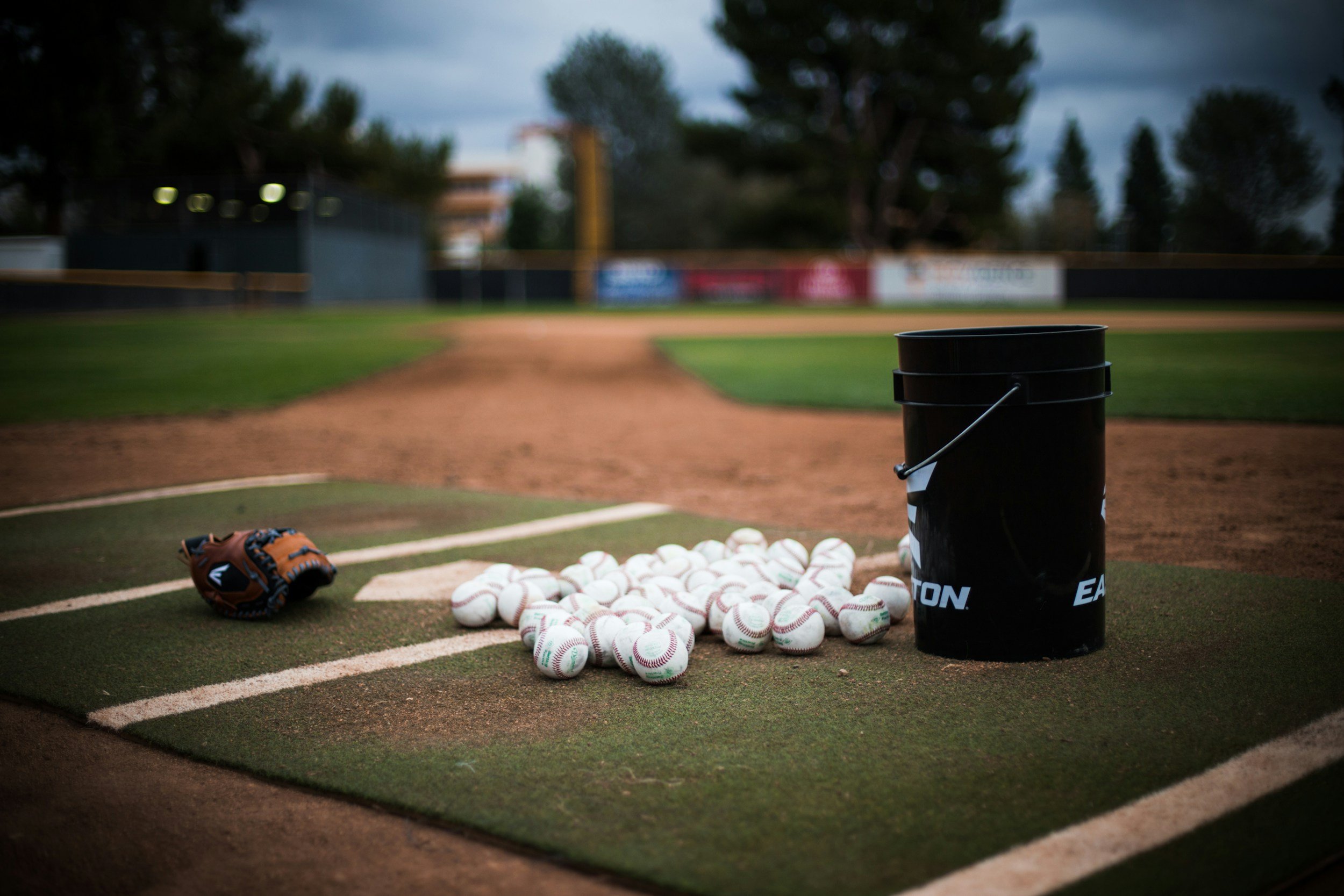 A baseball field with a mound of baseballs, a black bucket, and a glove on home plate.