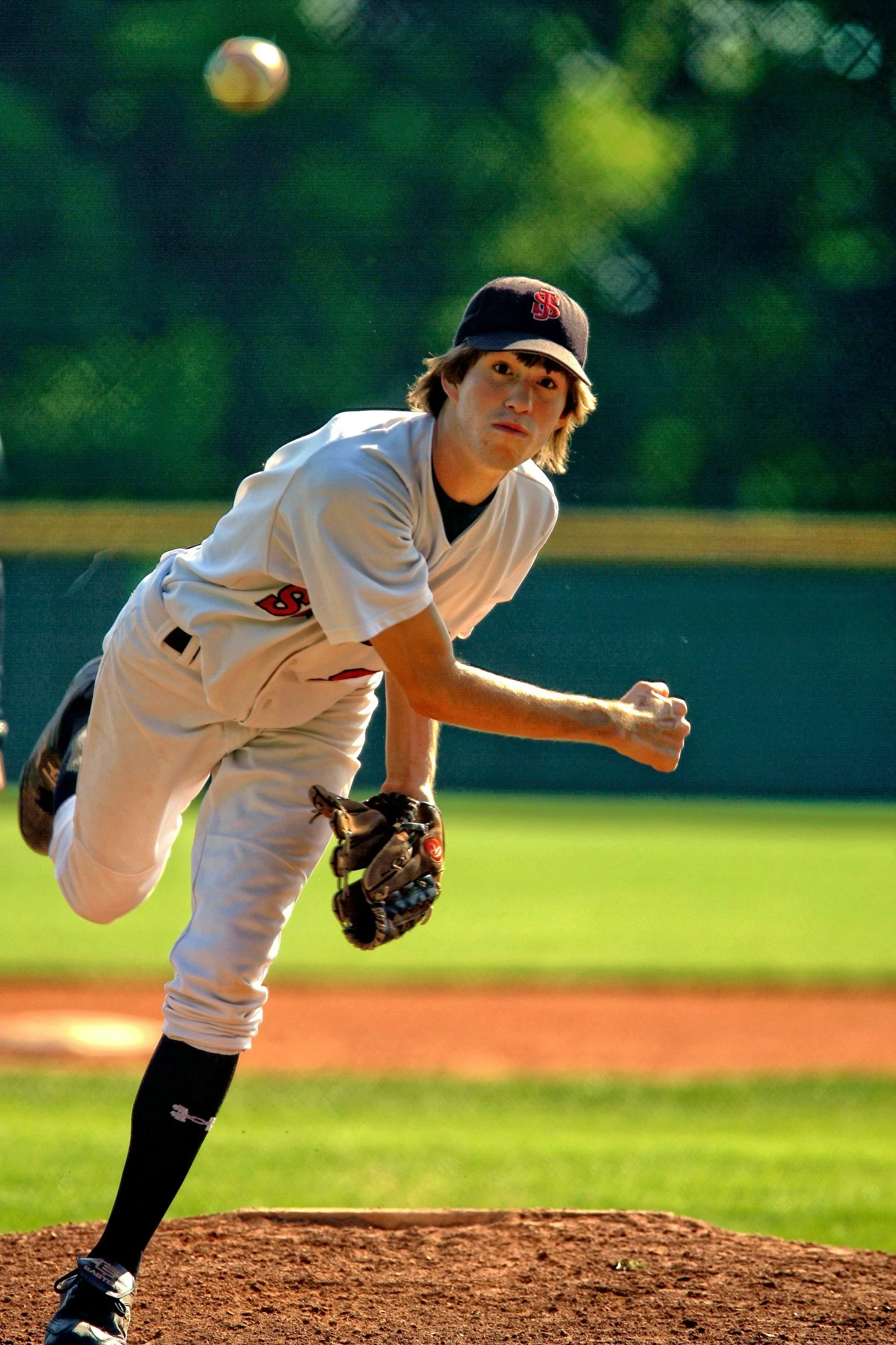 A baseball pitcher in uniform and cap throws a pitch on the mound, with a baseball in the air above him on a grassy baseball field.