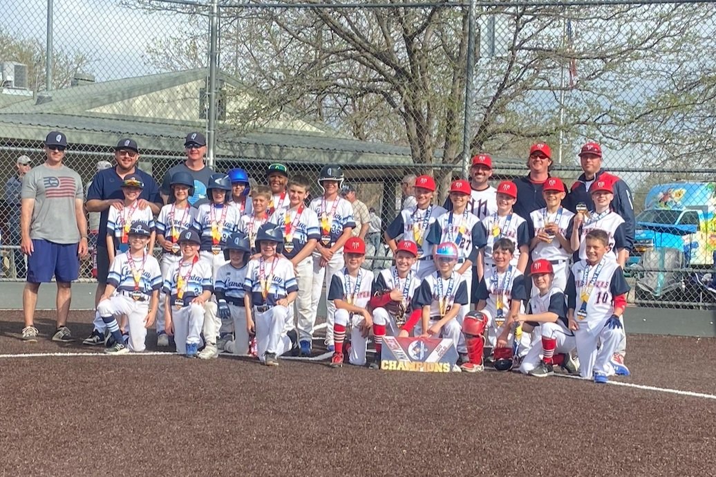 Youth baseball team wearing medals, posing on a baseball field with coaches, holding a champion banner.
