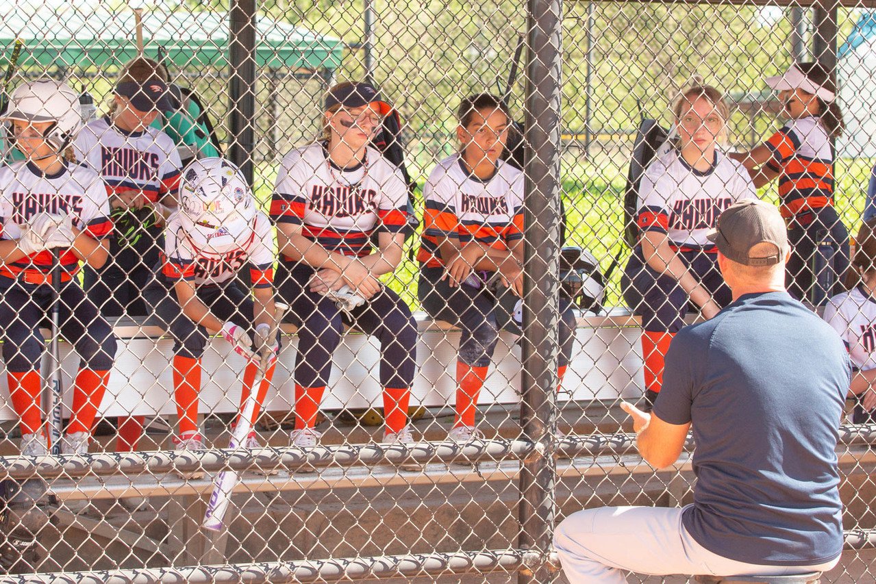 A youth baseball coach speaking to a team of young players sitting on a bench behind a chain-link fence.