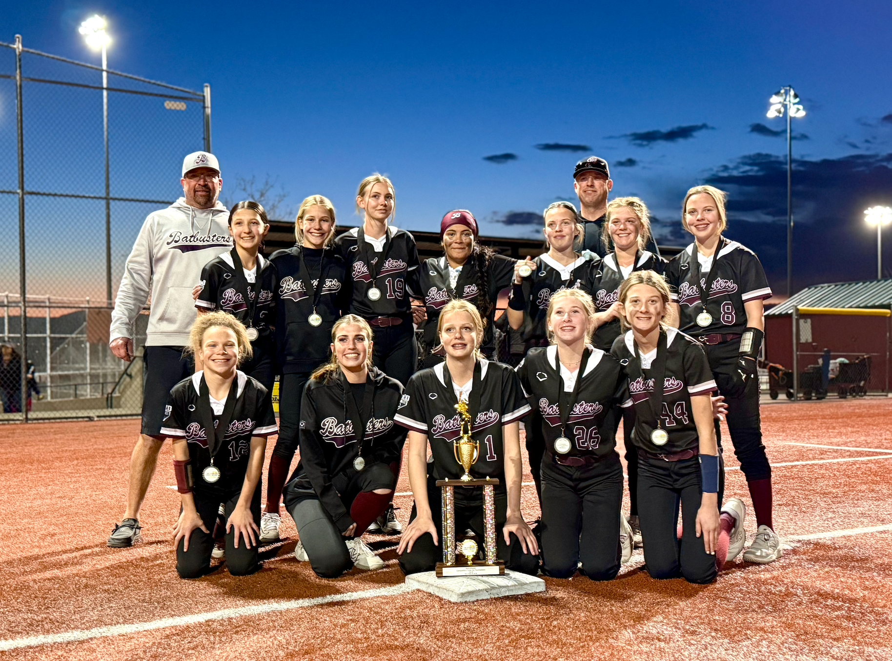 Youth softball team in black uniforms with medals, posing with a trophy on a softball field at dusk. Two coaches stand behind the team, one in a white hoodie and cap. The team celebrates a victory.
