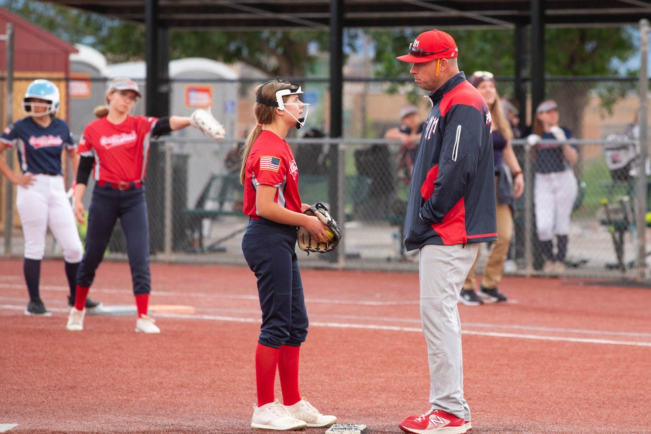 A young girl in a softball uniform holding a glove stands on a softball field, talking to a coach. The coach is listening with his hands in his pockets. In the background, other players and spectators are visible near a chain-link fence.