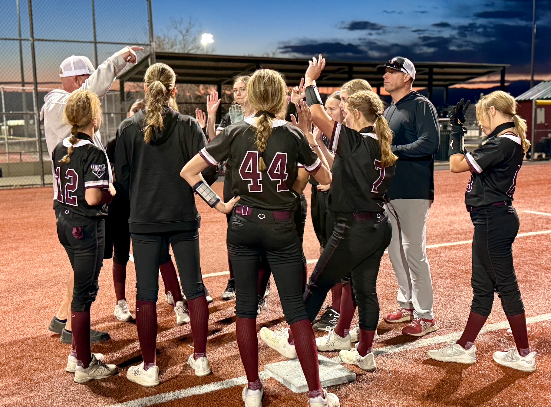 A nighttime softball team huddle on a field with players wearing black uniforms and coaches speaking to them.