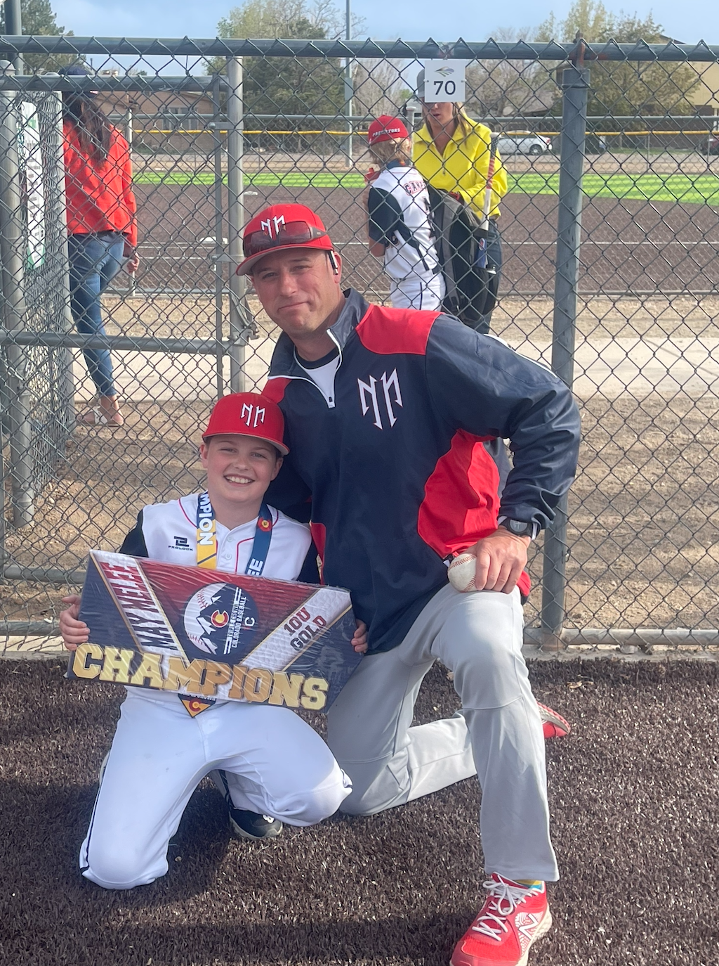 A young baseball player and coach kneeling on a baseball field, posing with a championship sign that says 'Champions' and a medal. The player is wearing a white uniform and red cap, and the coach is wearing a dark jacket with red and white accents an