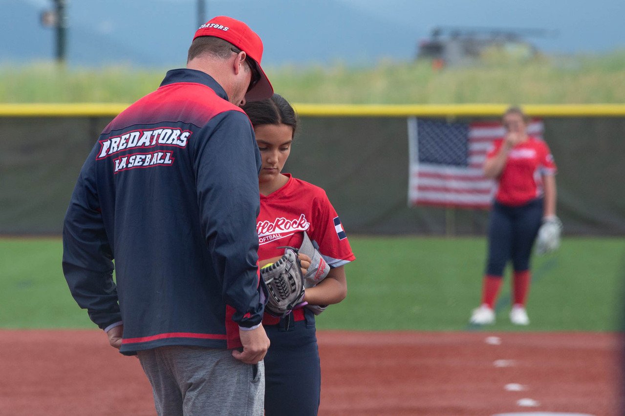 A baseball coach and a young female player are standing on the field during a game, with a referee standing in the background near an American flag. The coach is wearing a navy and red jacket that says 'PREDATORS BASEBALL' on the back, and the player