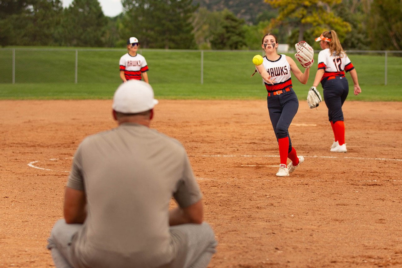 A girl in a softball uniform catching a yellow softball with her glove on a softball field, with two other players and a man sitting behind her.