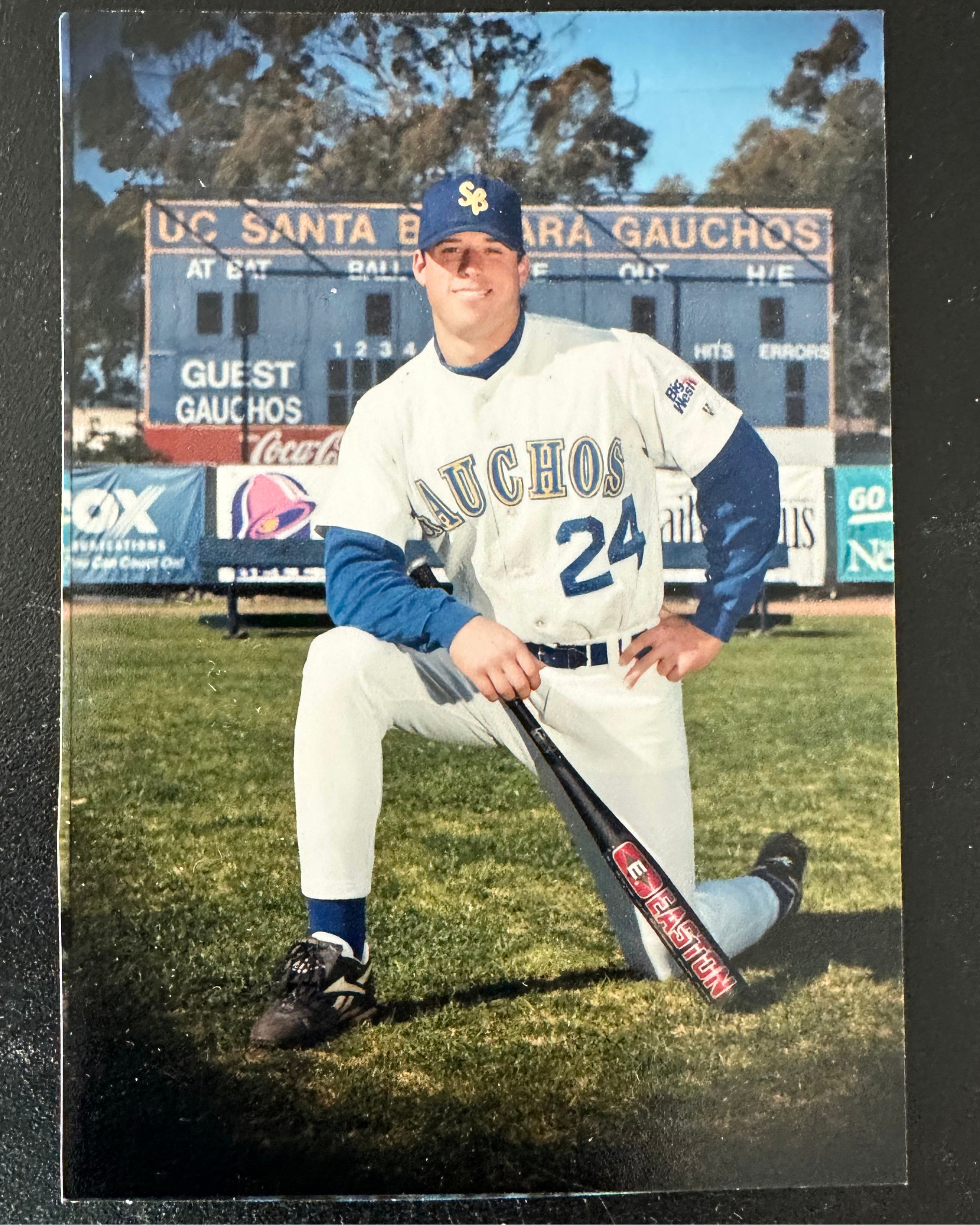 A young baseball player in a white and blue UC Gauchos uniform with the number 24, posing on the field with one knee on the grass, holding a bat, in front of a scoreboard.