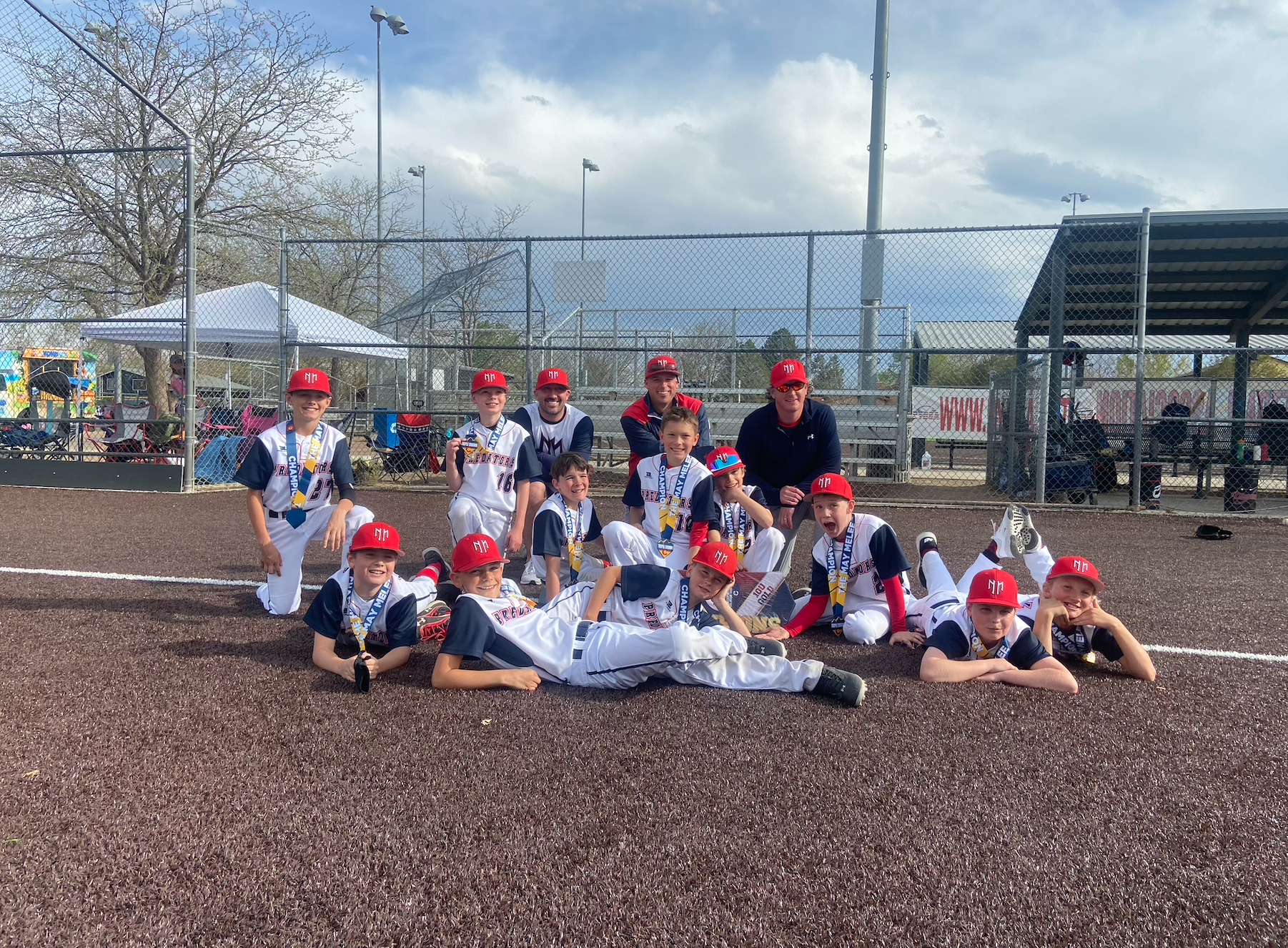 A youth baseball team in uniform with medals around their necks, some kneeling and lying on the ground, posing for a group photo on a baseball field with a fenced background and cloudy sky.