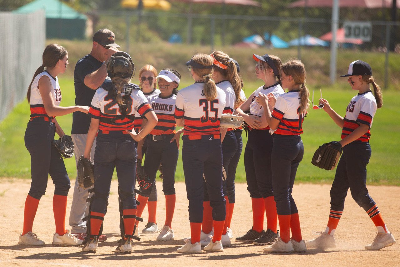 A group of young female softball players in white, black, and red uniforms gathered around their coach on a softball field.