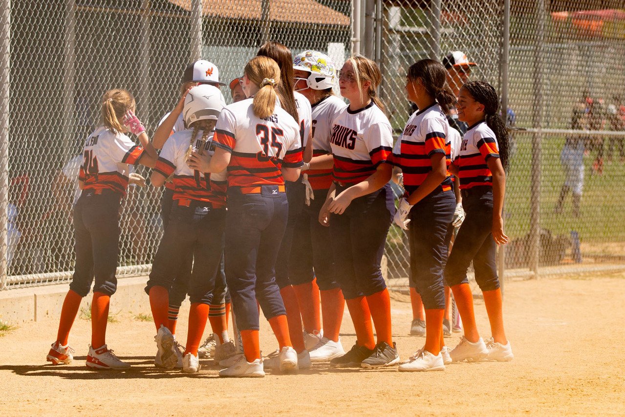 Girls baseball team gathered on the field during a game, wearing uniforms with black, orange, and white colors, some wearing helmets and gloves.