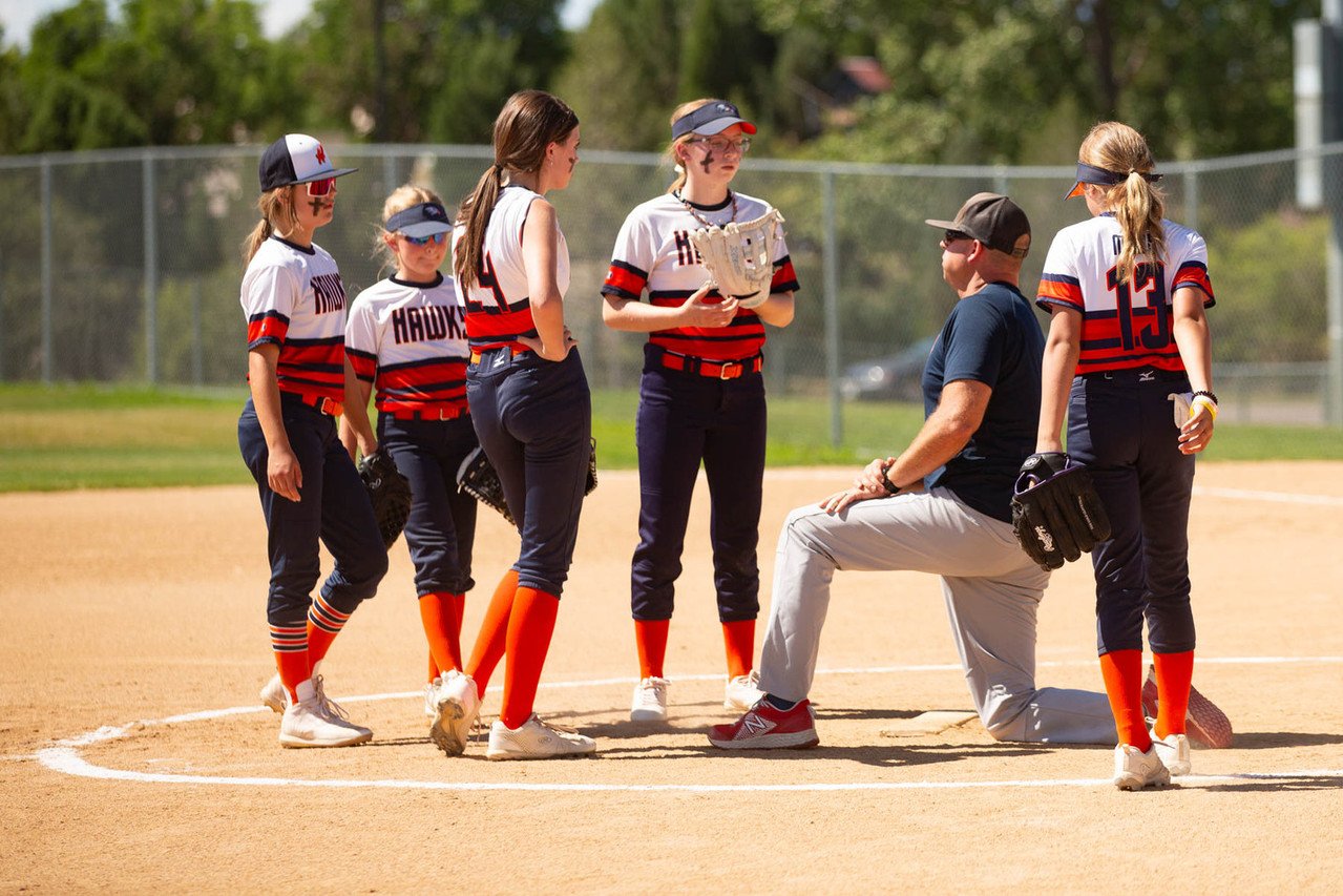 Softball coach kneeling and talking to young female players on the field.