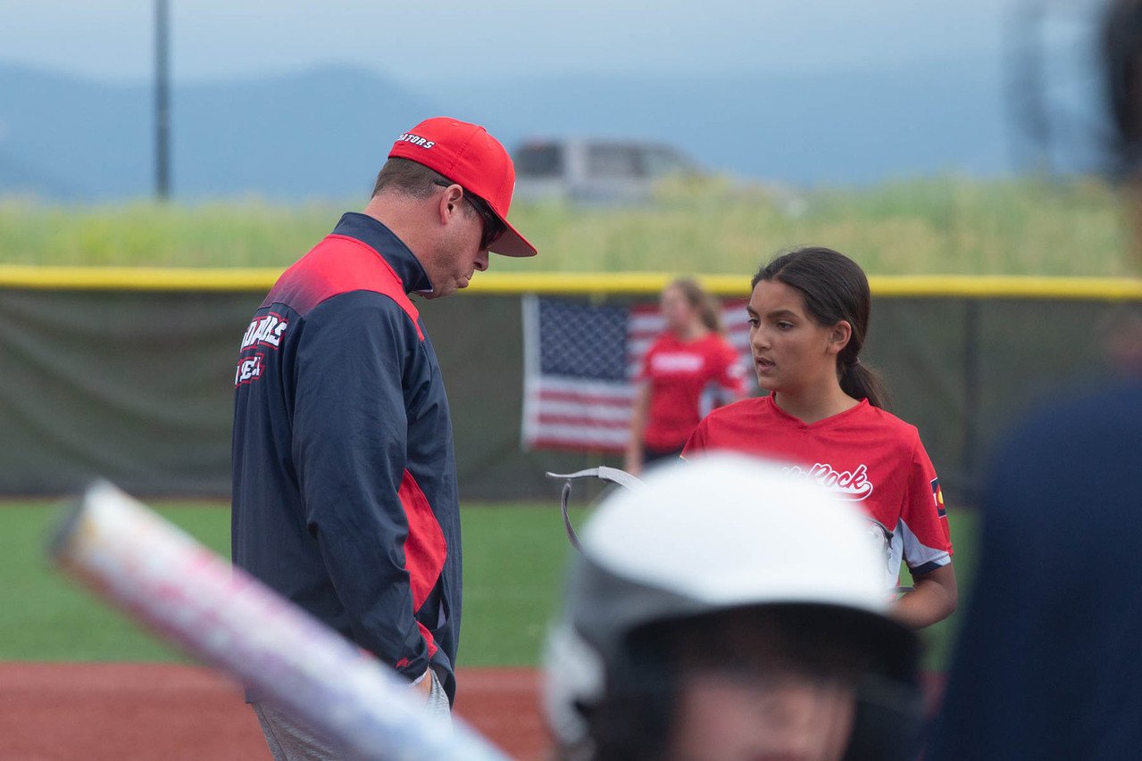 A young girl and a man in baseball uniforms standing on a baseball field with an American flag in the background.