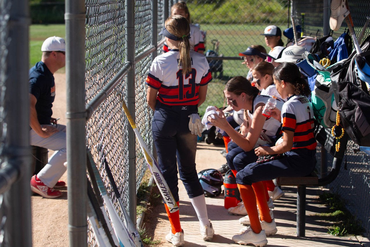 Girls' softball team sitting on a dugout bench, some preparing to play while a coach stands nearby during daytime