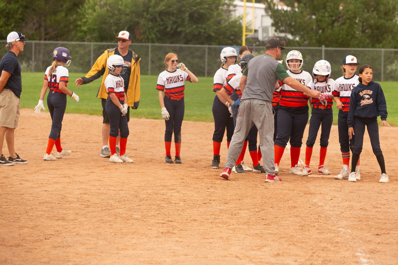 Youth girls' softball team and coaches on a field, with some players wearing helmets and team uniforms, during a practice or game.