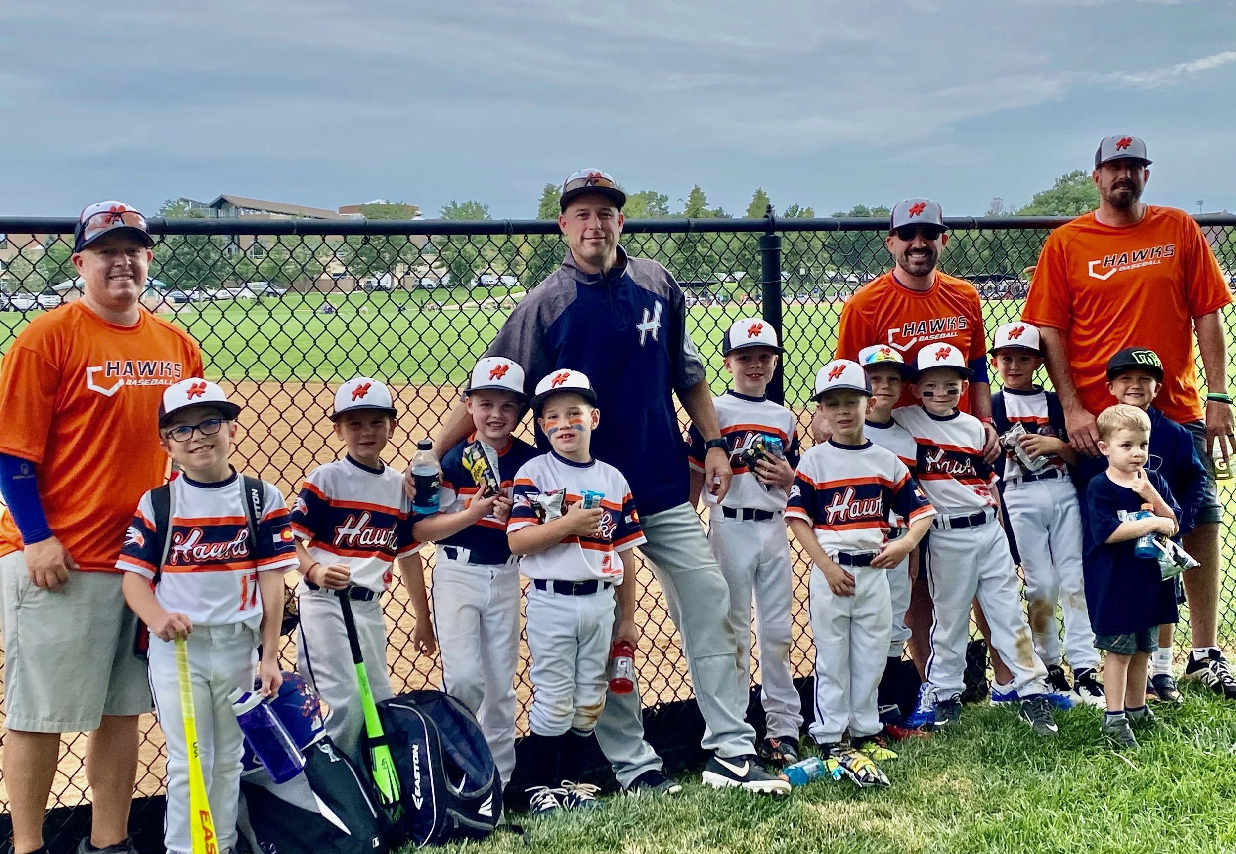 A youth baseball team with coaches standing on the grass in front of a baseball field, wearing team uniforms and caps.