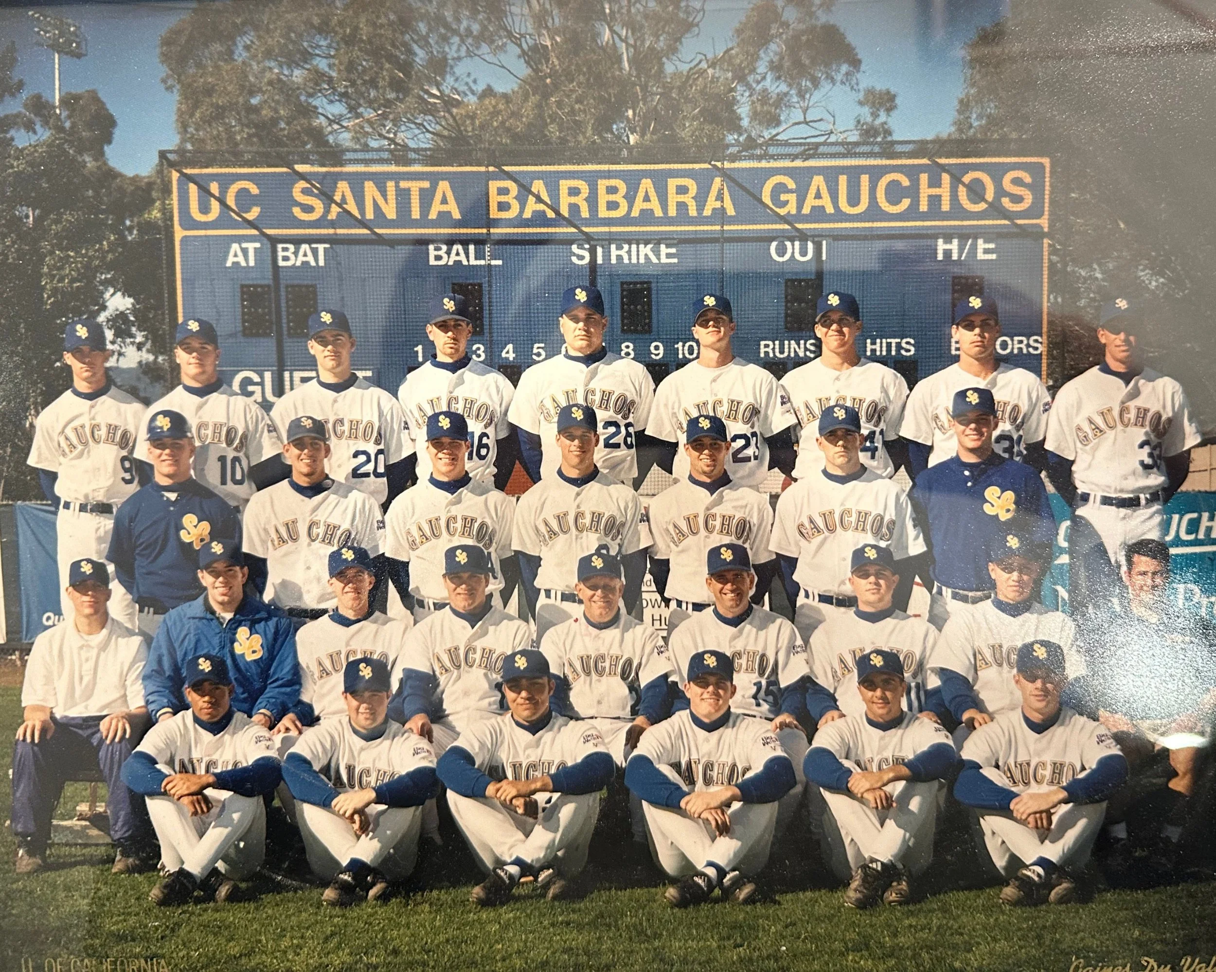 A group of baseball players and coaches posing for a team photo in front of a scoreboard at UC Santa Barbara Gauchos, dressed in white and blue uniforms with team logos and caps, outdoors on a sunny day.