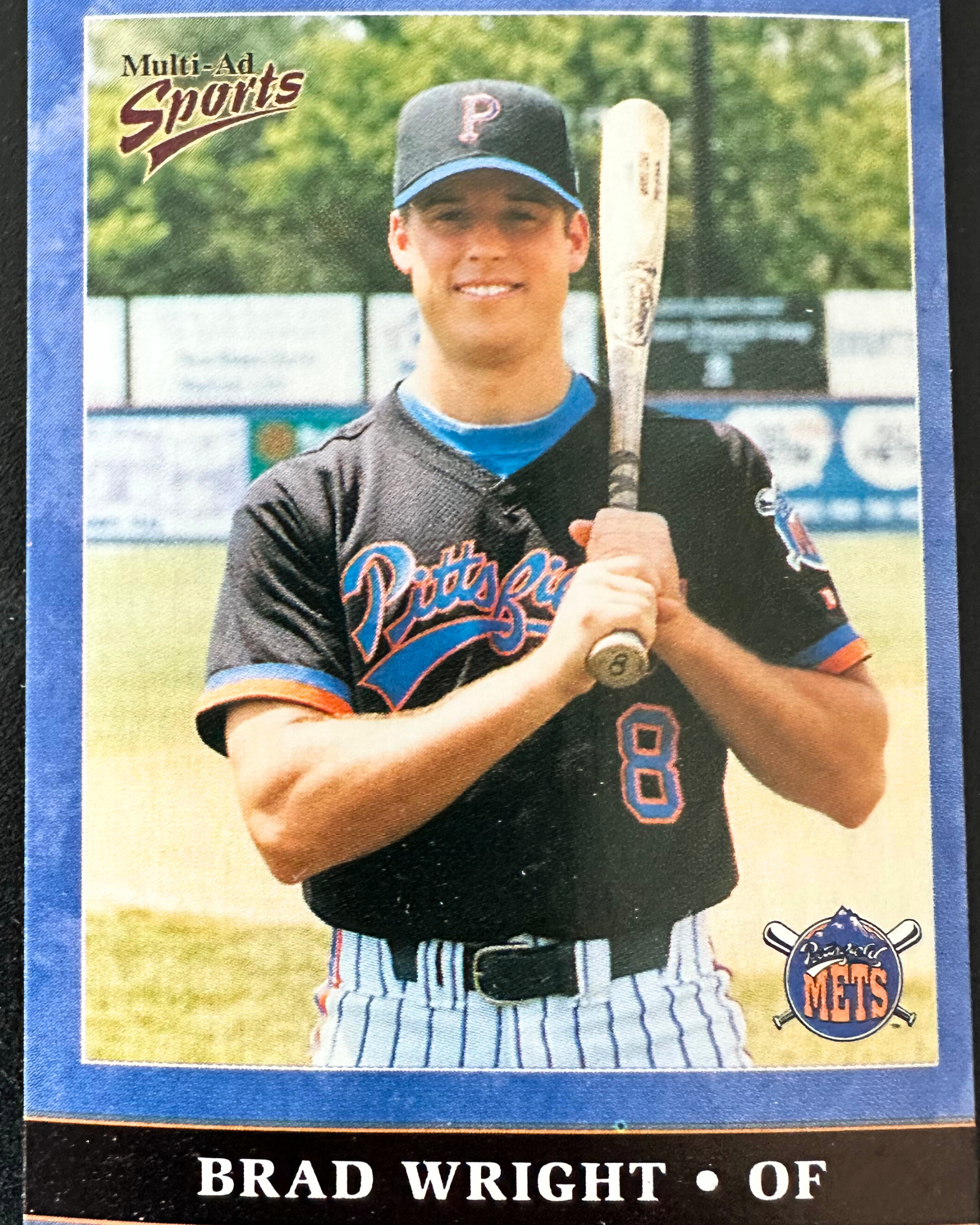 Photograph of a young baseball player wearing a Pittsburgh Mets uniform, holding a bat, and standing on a baseball field with a fence and trees in the background.