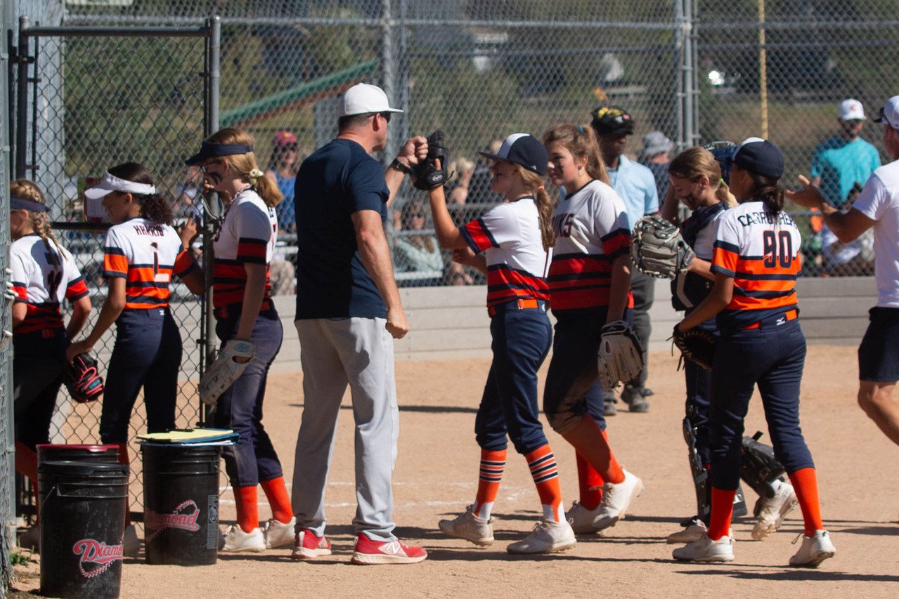 Young female softball players and coaches gathered on a dirt field, some with gloves, near a chain-link fence.