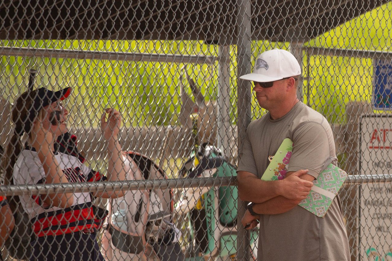 A man wearing a white cap, sunglasses, and a gray T-shirt stands outside a chain-link fence, holding a notebook and a folder, watching a girl in a racing uniform who is talking to someone inside the fenced area.