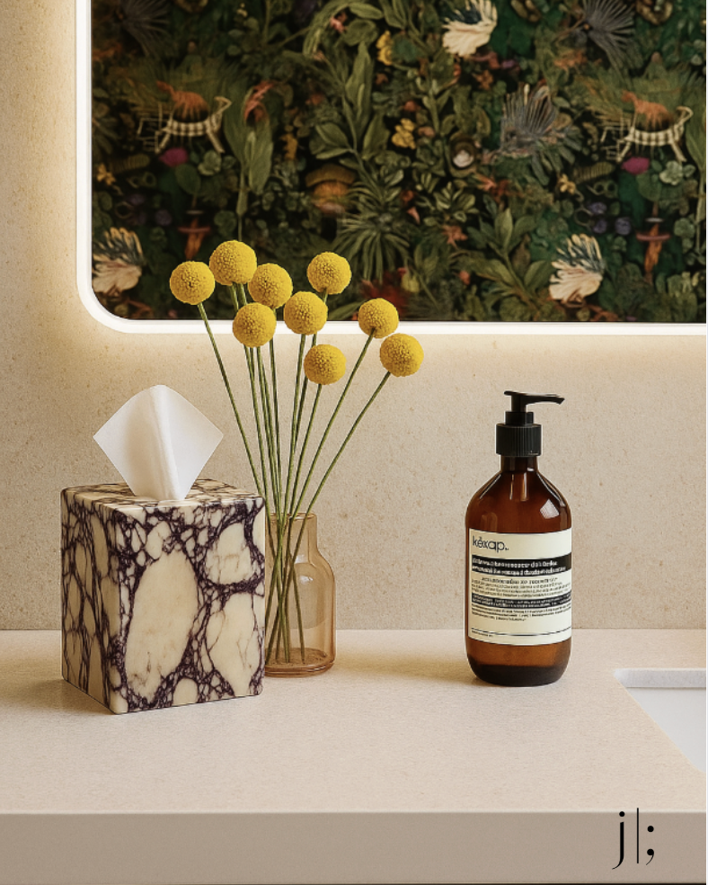A bathroom countertop with a marble tissue box, a vase with yellow billy ball flowers, a brown soap dispenser, and a mirror reflecting a green and floral wallpaper.