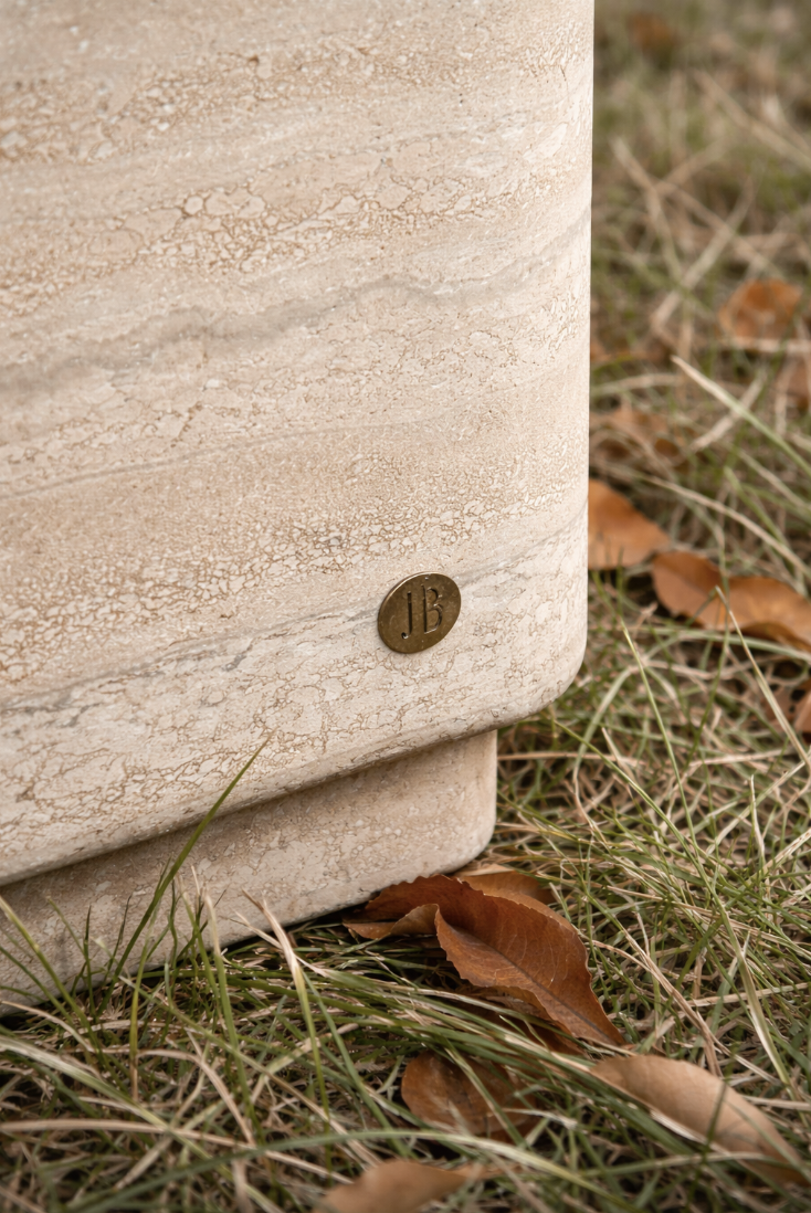 Close-up of a beige marble-like surface with a small bronze-colored metal tag labeled 'JB' attached near the bottom edge, surrounded by grass and fallen brown leaves.