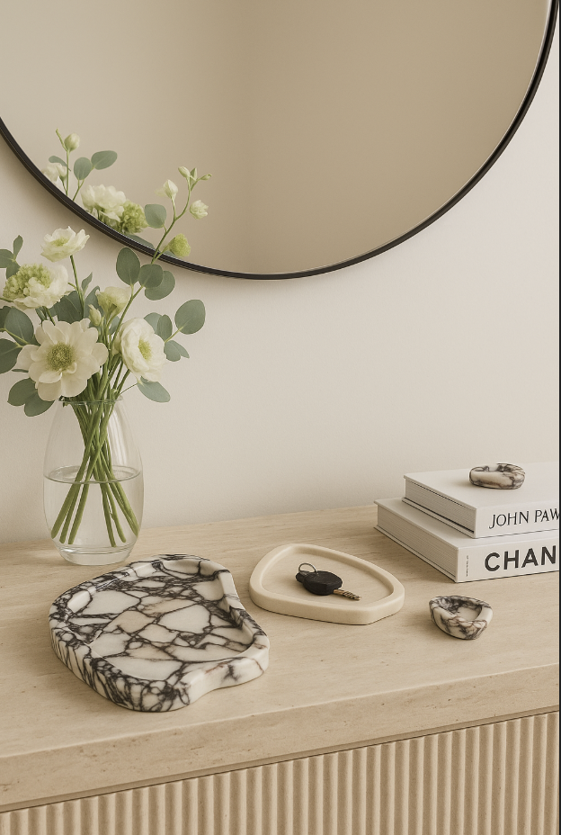 Decorative tabletop with a vase of white flowers, a round mirror on the wall, black and white marble tray, beige ceramic dish with a key, and a small stone bowl.